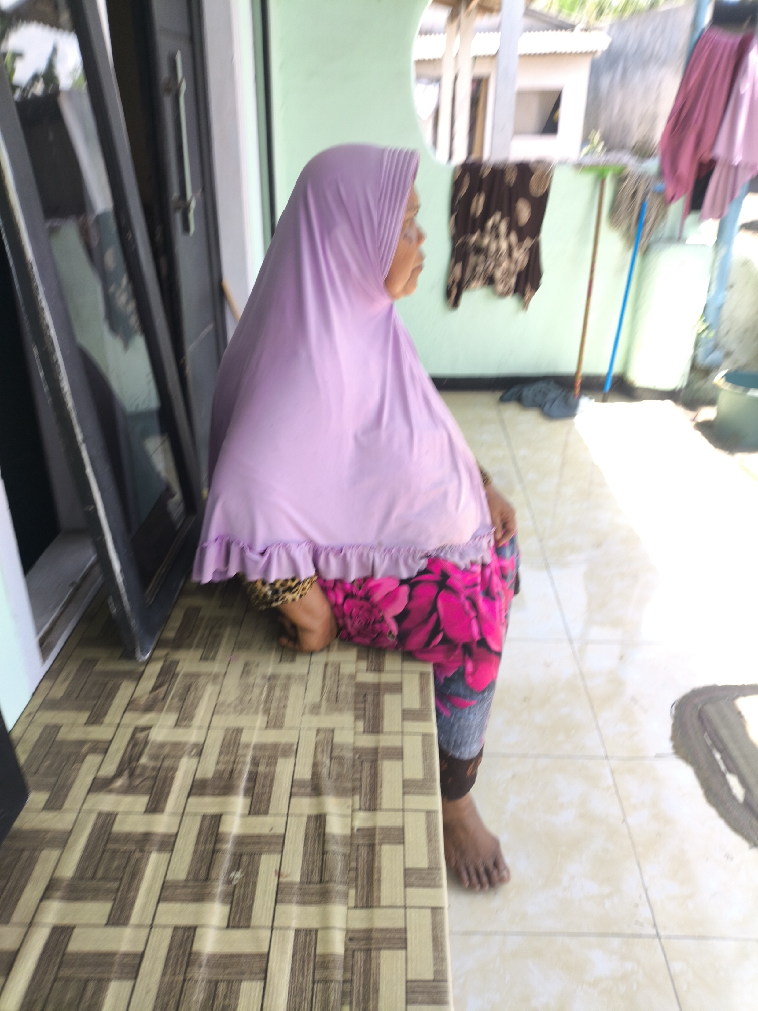 In the city of Grati, Indonesia, an Indonesian woman wearing a light purple hijab and a patterned skirt is seated on a bench. The bench has a tan and brown geometric pattern. To her left is a dark-colored door with a silver handle. Behind her, a light green wall is visible, with a brown and tan patterned cloth hanging on it. A mop with a blue handle leans against the wall. The ground is tiled with a light beige color. Sunlight illuminates the scene from the right, creating a bright glare on the floor.