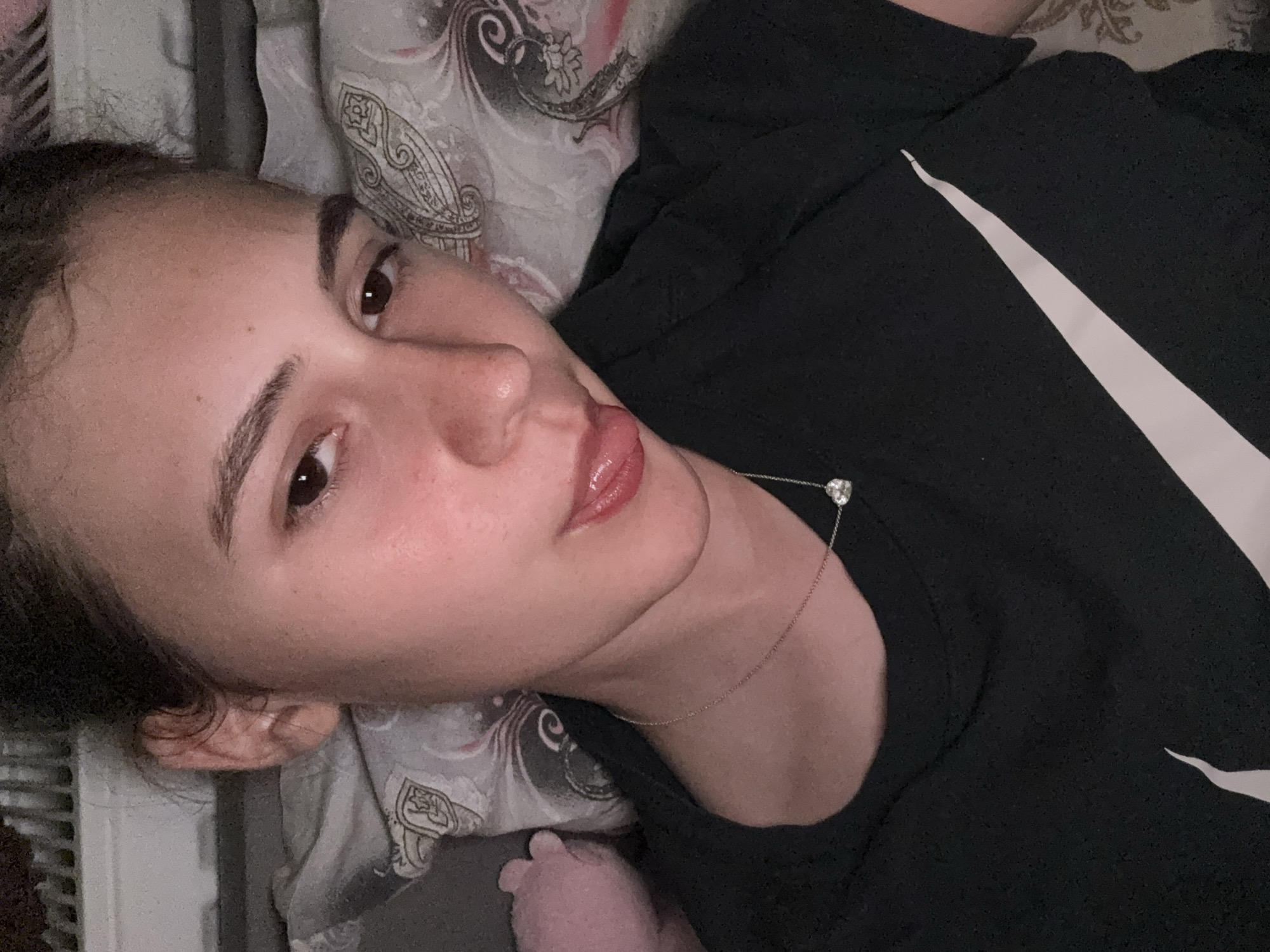 A young individual, appearing female, is centered in the frame, lying down on what appears to be a bed, with her head resting on a patterned pillow. She looks directly into the camera with dark eyes, her tongue slightly extended from her mouth. Her hair is dark and pulled back, with some loose strands visible near her right ear. Her skin is fair with visible freckles. She wears a black short-sleeved t-shirt featuring a large white Nike swoosh logo across the chest. Around her neck is a delicate silver-colored chain necklace with a small, clear, heart-shaped pendant. The pillow behind her head is light-colored with a dark, intricate, paisley-like pattern. To her left, a soft pink object, possibly a portion of a stuffed animal, is partially visible. In the background, behind her head, a light-colored, ridged structure, resembling a radiator or blinds, can be observed. The specific location in the city of Panagyurishte, Bulgaria, cannot be determined from the visual information provided in the image.