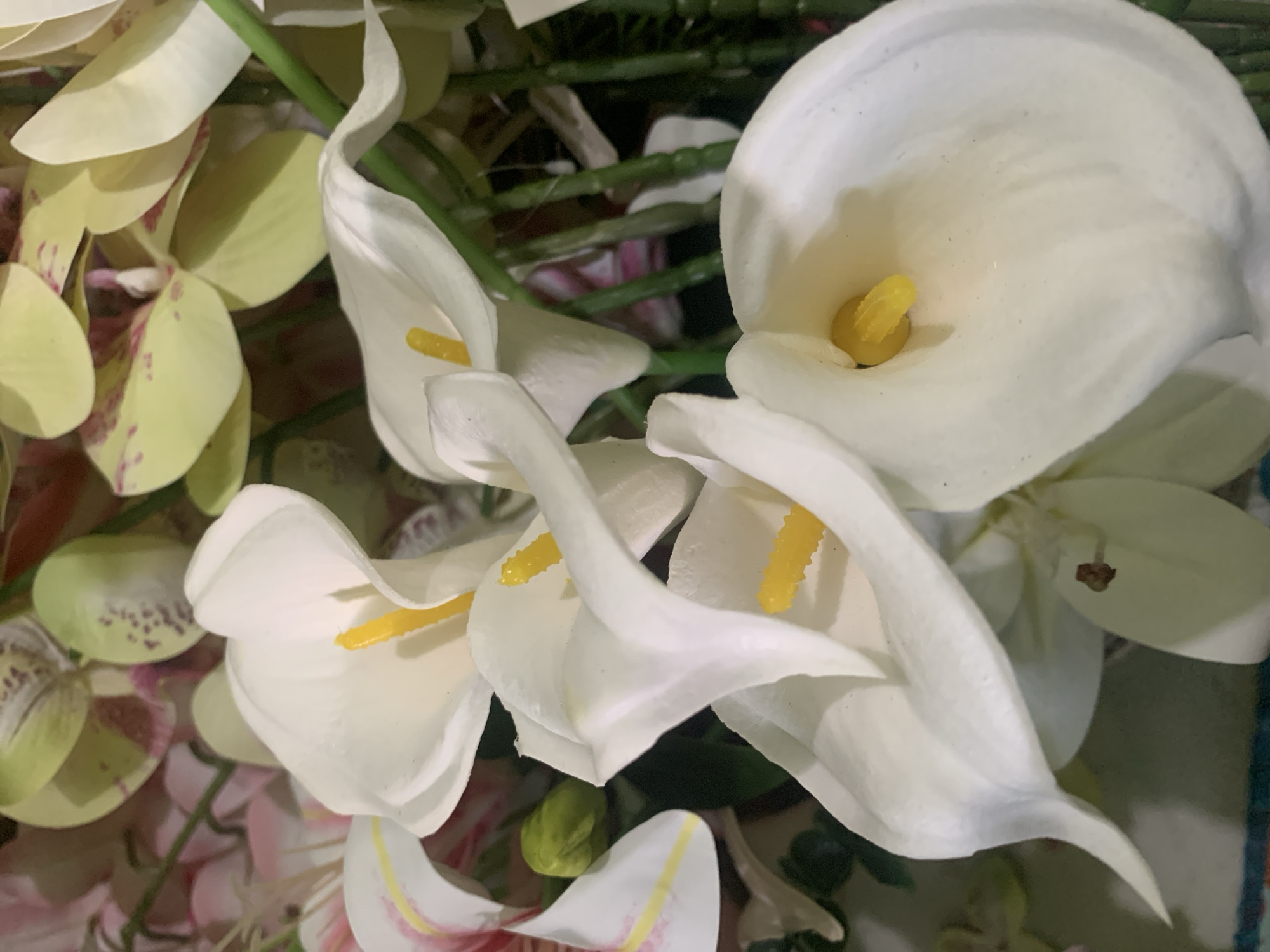 The image shows a close-up view of a bouquet of white calla lilies and other flowers. The calla lilies are the dominant subject, with their distinctive trumpet shape and yellow spadix. Some of the lilies are in full bloom, while others are partially open. The background is filled with other flowers, including what appear to be pink orchids and possibly some greenery. The lighting suggests an indoor setting, possibly a floral arrangement for decoration. The image does not contain any information that could identify its location within Kuala Pembuang, Indonesia.