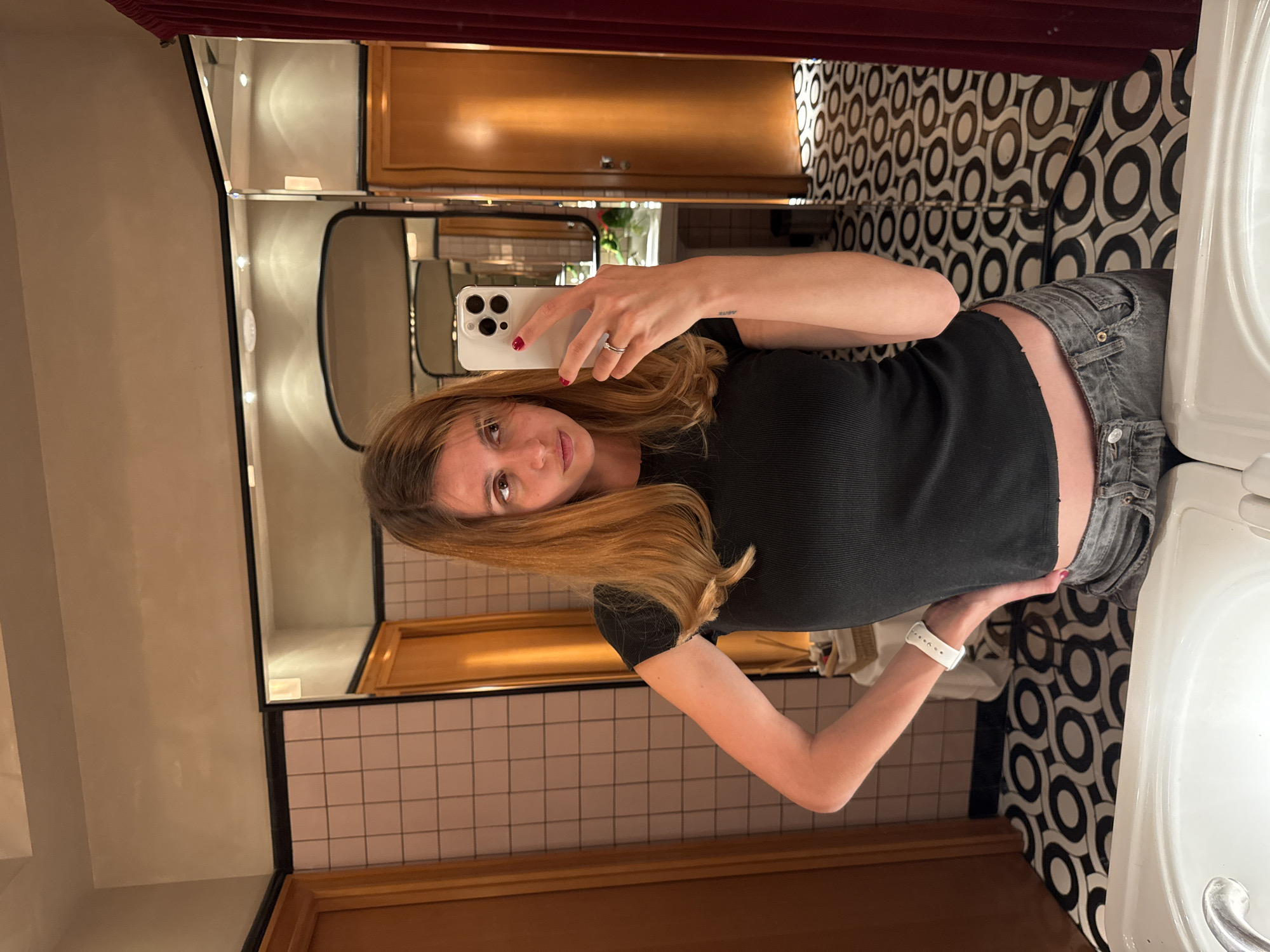 A woman is taking a selfie in a bathroom. The bathroom features checkered black and white floor tiles and pinkish-beige wall tiles with black grout. The countertop surrounding the sink is white. There is a large mirror in front of her, reflecting the bathroom's interior. There are cabinets with wooden fronts above the mirror and pink curtains at the top of the frame. The woman is wearing a black top and grey shorts. She holds a white smartphone in her right hand, taking the picture. Her hair is blonde, and she's wearing a white smartwatch on her left wrist. The location is Yerevan, Armenia.