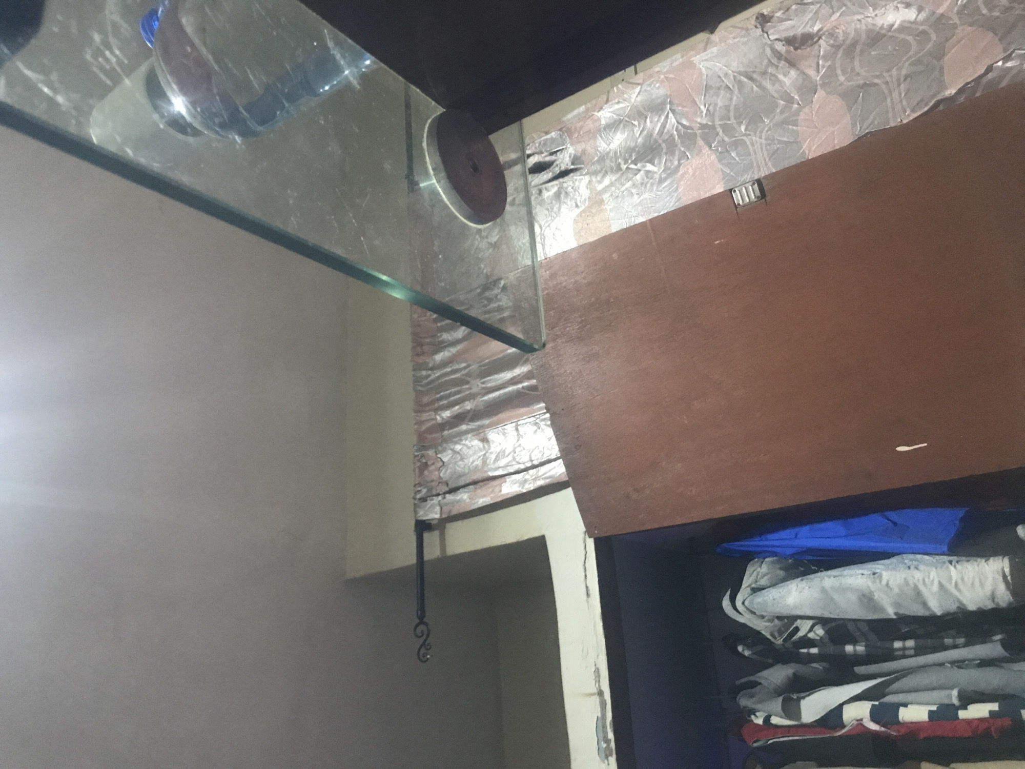 The image depicts an interior scene, likely a closet or wardrobe, and an adjacent area with a glass shelf. The closet contains several items of clothing hanging on a rod, including jackets and shirts in various colors and patterns. One item of clothing is a bright blue garment. The closet doors appear to be made of a dark reddish-brown material.
Above the closet, there is a curtain with a patterned design, possibly floral, in shades of beige and pink. A decorative metal rod supports the curtain.
In the background, to the right of the closet, is a glass shelf or surface, angled upwards. On this shelf, a clear plastic bottle with a blue cap is visible. Below the glass, there is a dark circular object with a hole in the center, possibly a weight or decorative element. The lighting in the scene is dim, with a bright light source emanating from the upper left, casting shadows.
The location is described as Kabong, Nigeria. However, there are no specific visual cues in the image that definitively place it within the city of Kabong, Nigeria. The scene appears to be a domestic interior setting.