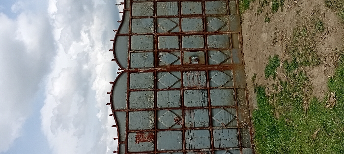 A two-panel, double-leaf metal gate, exhibiting significant rust and peeling light blue-grey paint, stands centered in the frame in Heni Village, Kenya. The gate features a grid construction composed of primarily rectangular and central diamond-shaped panels. Each half of the gate is topped by a distinct arched section with multiple small, pointed finials. The ground immediately in front of the gate consists of a mixed surface of sparse green grass and exposed brown dirt. Above the gate, the sky is partially overcast with a mix of white and grey cumulus clouds. No individuals, animals, or active events are present within the scene.