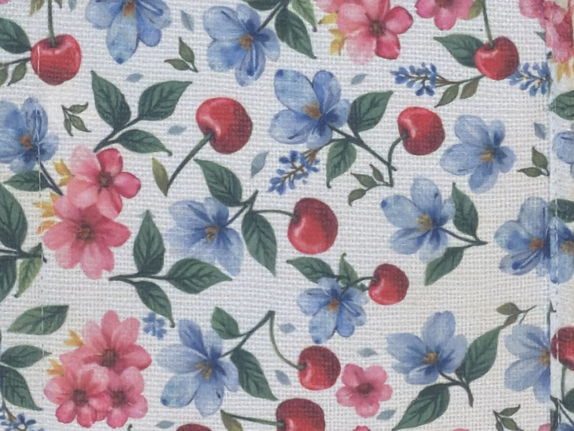 This is a close-up, top-down, eye-level shot of a fabric with a watercolor floral pattern. The pattern features red cherries, pink and blue flowers, and green leaves scattered across a textured, light-colored background. The style is illustrative and appears to be a repeating pattern. No people, specific setting, time of day, weather, or emotions are depicted. There is no visible text.