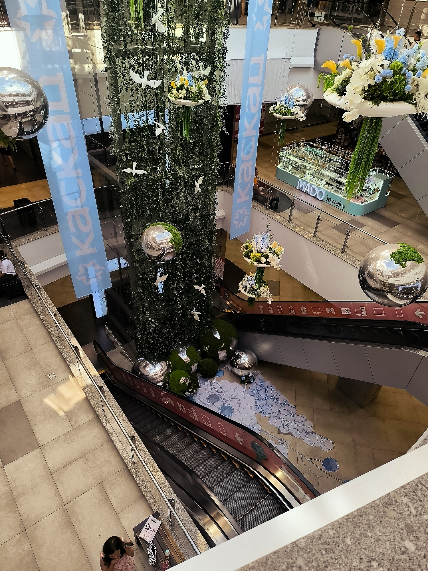 The image shows an interior view of a multi-level shopping mall, likely in Qarağandı city administration, Kazakhstan. The perspective is from a higher floor looking down. A large, green, hanging structure, possibly a living plant wall, dominates the center, adorned with white bird cutouts and some floral arrangements. Large, mirrored spheres are suspended around the central structure and also appear on lower levels, reflecting the surroundings.
To the left, a light blue banner with white lettering and star patterns hangs vertically. Below this banner, a person in dark clothing is seated on a bench. Further down, an escalator is visible.
In the mid-ground, a display case with the text "MADO Jewelry" is visible, lit from within. Another escalator is present in the foreground, with a patterned floor decoration featuring stylized blue flowers beneath it.
On the bottom floor, near the escalator, a woman in a pink dress is standing, looking down at something in her hands or on a surface in front of her. She is near a bench with some items on it, including a bag and what appears to be a shopping bag.
The overall environment is clean and modern, with a focus on decorative elements that suggest a festive or well-maintained atmosphere. The lighting is bright, indicating ample indoor illumination.