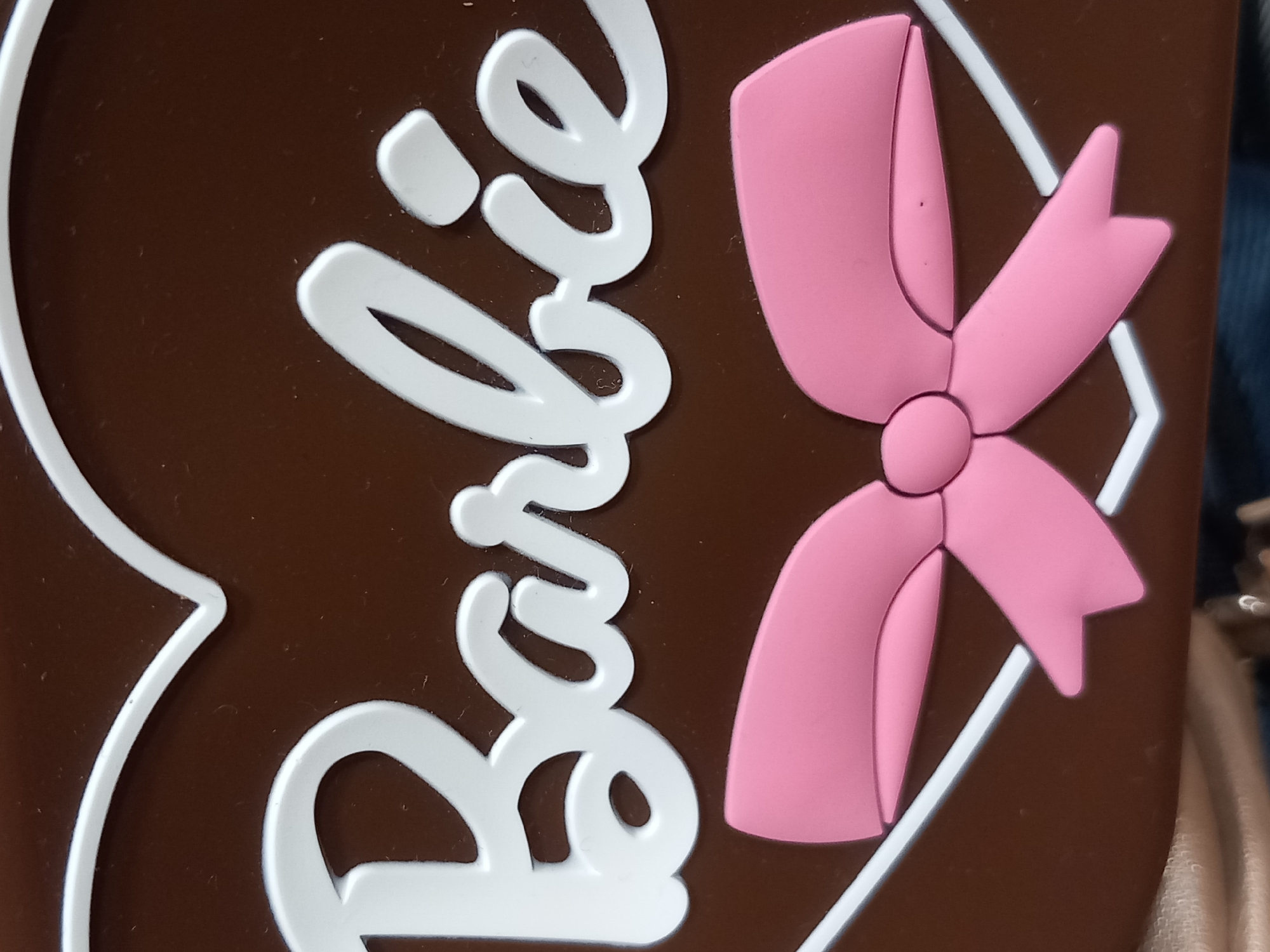The image is a close-up shot of a brown surface with white and pink raised lettering and a pink bow. The white lettering spells out "Barbie" in a cursive font, with the letters appearing to be made of a raised material. To the right of the text is a large, stylized pink bow with five loops. The bow also appears to be made of a raised material. The background is a solid brown. There are no people or any indication of a specific scene or setting other than the surface itself. No activity or event is taking place. The image is well-lit, and there are no discernible time of day, weather, or emotional cues. The only visible text is the word "Barbie".