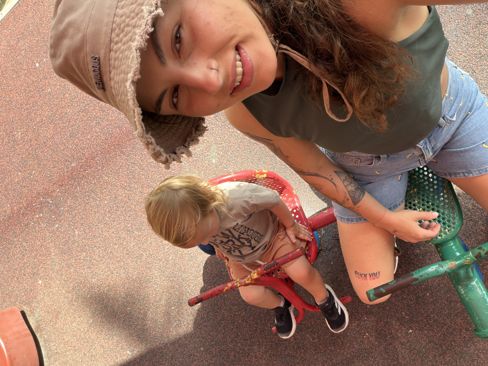 A woman and a child are positioned on a playground in Kiryat Motzkin, Israel. The woman, wearing a tan bucket hat, dark green tank top, and denim shorts with banana designs, is smiling at the camera. She has a tattoo on her arm and another tattoo on her right thigh that reads "FUCK YOU." The child, with blonde hair, is seated on a red playground ride, wearing a tan shirt with dark text, and black sneakers with white stripes. The playground surface is red. A green playground apparatus is visible next to the woman.