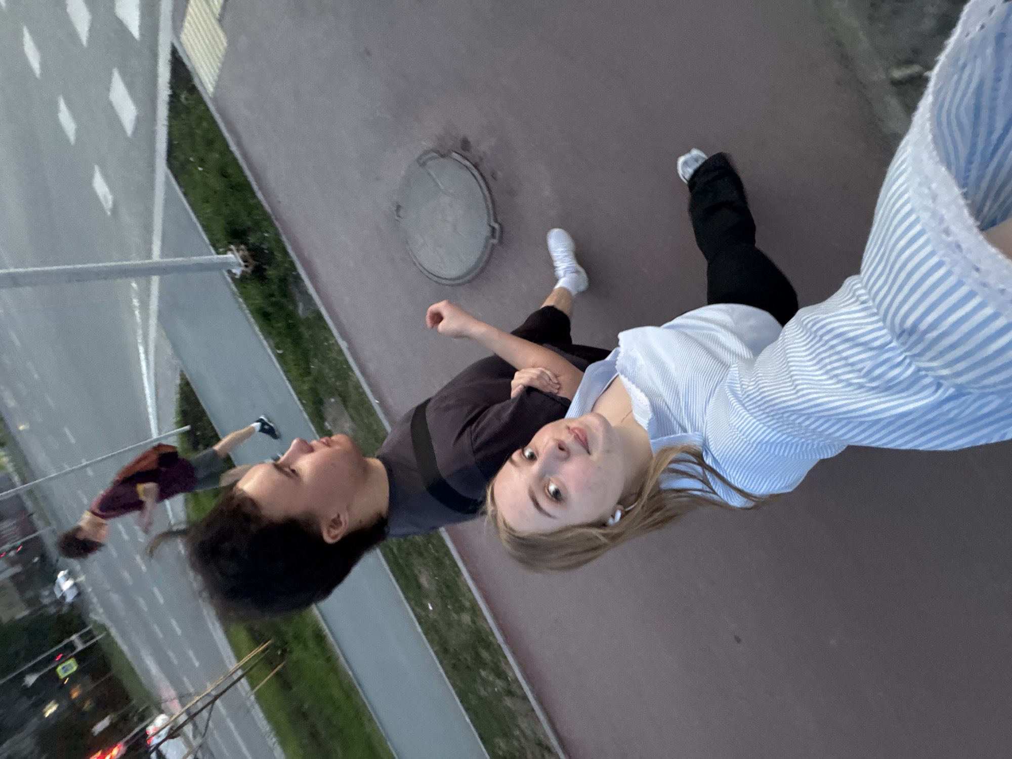 The photograph shows two individuals standing on a sidewalk alongside a bridge or overpass, likely in the city of Perm, Russia. One individual, seen in profile, has dark hair and is wearing a dark gray or black t-shirt. The other person, positioned closer to the camera, is wearing a light blue and white striped shirt and has blonde hair. They are standing next to a manhole cover on the sidewalk. Beyond the sidewalk, there is a grassy area and a road below with vehicle traffic. A pedestrian bridge is also visible in the background.