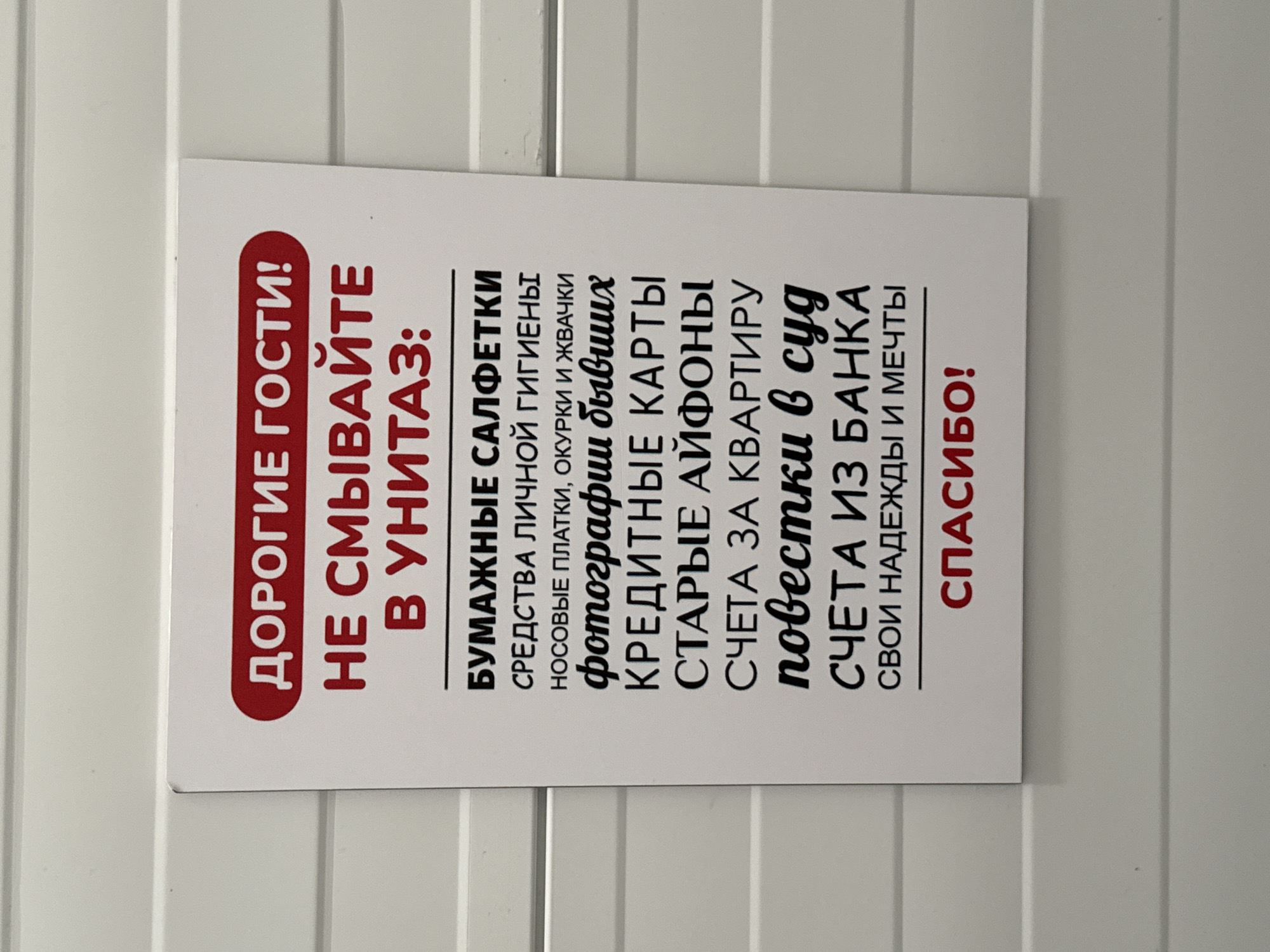 A close-up, eye-level, full-frame, outdoor, daylight, medium shot shows a white sign with red and black text, mounted on a horizontally ribbed white surface. The sign is centrally located. The text is in Russian. The upper portion of the sign has a red oval with white text that reads "ДОРОГИЕ ГОСТИ!" (Dear Guests!). Below this, in large red block letters, is "НЕ СМЫВАЙТЕ В УНИТАЗ:" (Do not flush in the toilet:). The remainder of the sign lists items that should not be flushed, with each item on a new line. The items are: "БУМАЖНЫЕ САЛФЕТКИ" (paper napkins), "СРЕДСТВА ЛИЧНОЙ ГИГИЕНЫ" (personal hygiene products), "НОСОВЫЕ ПЛАТКИ, ОКУРКИ И ЖВАЧКИ" (tissues, cigarette butts, and chewing gum), "фотографии бывших" (photos of ex-partners), "КРЕДИТНЫЕ КАРТЫ" (credit cards), "СТАРЫЕ АЙФОНЫ" (old iPhones), "СЧЕТА ЗА КВАРТИРУ" (apartment bills), "повестки в суд" (court summons), "СЧЕТА ИЗ БАНКА" (bank statements), "СВОИ НАДЕЖДЫ И МЕЧТЫ" (your hopes and dreams). The sign concludes with a thin red line and below it, in red block letters, "СПАСИБО!" (Thank you!). The surface on which the sign is mounted appears to be an exterior wall of a building. The lighting suggests daylight. The image is sharp and clear.
