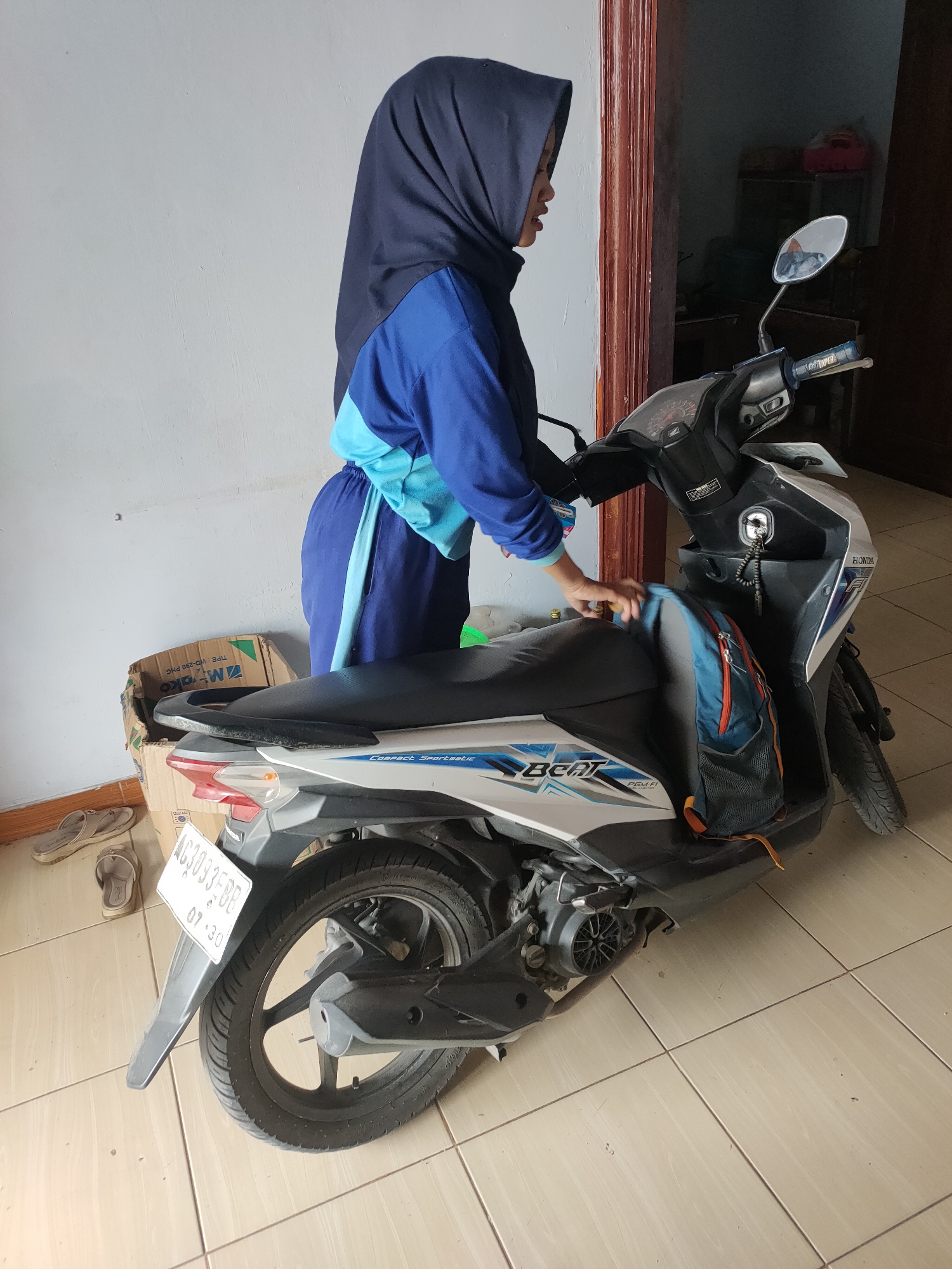 A young woman wearing a dark blue hijab and a blue and light blue tracksuit stands next to a white Honda Beat scooter. She is in profile, facing right, and her right hand is reaching towards the scooter's seat. A backpack with orange and black accents is attached to the back of the scooter. The scooter has a license plate that reads "AG 3093 BB". The background shows a light gray wall and a doorway with a brown frame. The floor is tiled with light beige square tiles. A cardboard box and a pair of flip-flops are visible on the left side of the image. The overall scene appears to be indoors, possibly in a home in Kanigoro, Indonesia, as indicated by the license plate.