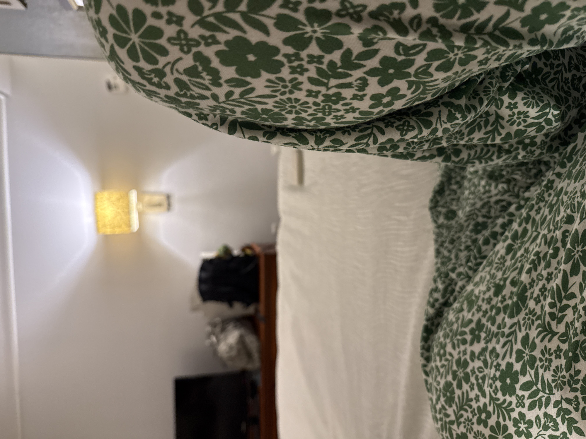 The image shows an interior scene. A white wall with a warm-toned wall sconce is visible on the left. In the background, there is a shelf or cabinet with several items, including a black bag. In the foreground, the image is dominated by a white and green floral patterned fabric, which appears to be clothing, possibly worn by the photographer. The location is inside a building in San Antonio, United States of America.