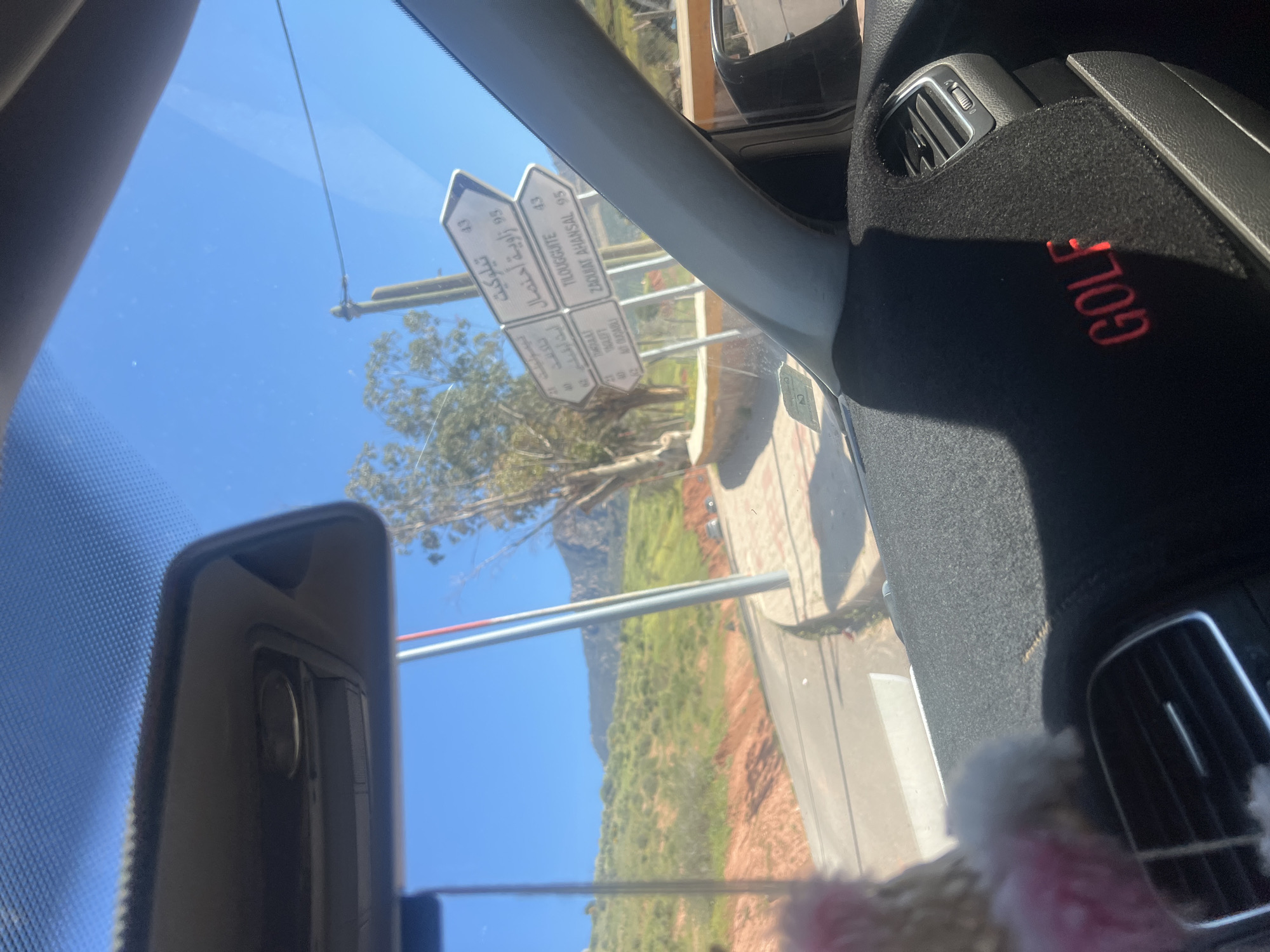 Captured through the windshield of a Golf brand vehicle, the image reveals a sunlit, outdoor scene in the city of Ouaouizert, Morocco. A road sign points to locations, reading: Tilougguite (43), Takaft (95), and Zaquiat Ahansal (95). The sign also references Moula and At Ouçabli. The landscape consists of a green, mountainous terrain, and a paved road. The car's interior features a black dashboard with the word "GOLF" embroidered in red, and air conditioning vents. A portion of the rearview mirror is visible. The sky is clear blue.