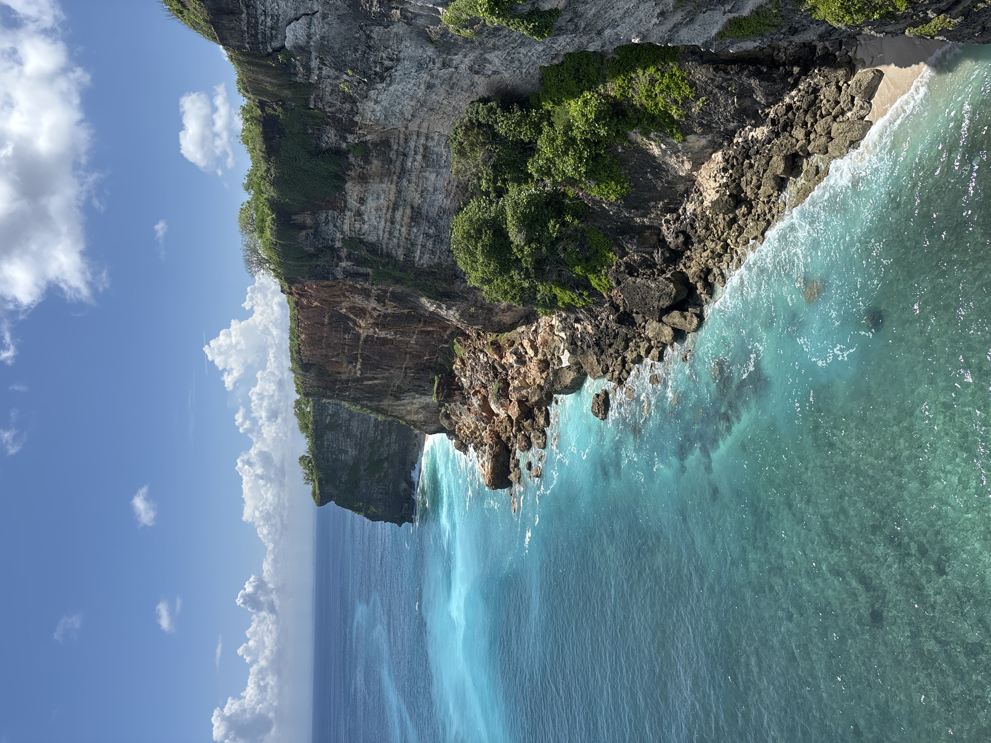 The image captures a coastal scene in Uluwatu, Indonesia, viewed from a high vantage point. A steep, rocky cliff dominates the landscape, with lush green vegetation clinging to its upper sections. The cliff face is textured with layers of rock and earth, descending into the turquoise waters of the ocean. The water is clear near the shore, revealing underwater rocks and the seabed, then deepens in color further out. The sky is a bright blue, scattered with puffy white clouds.