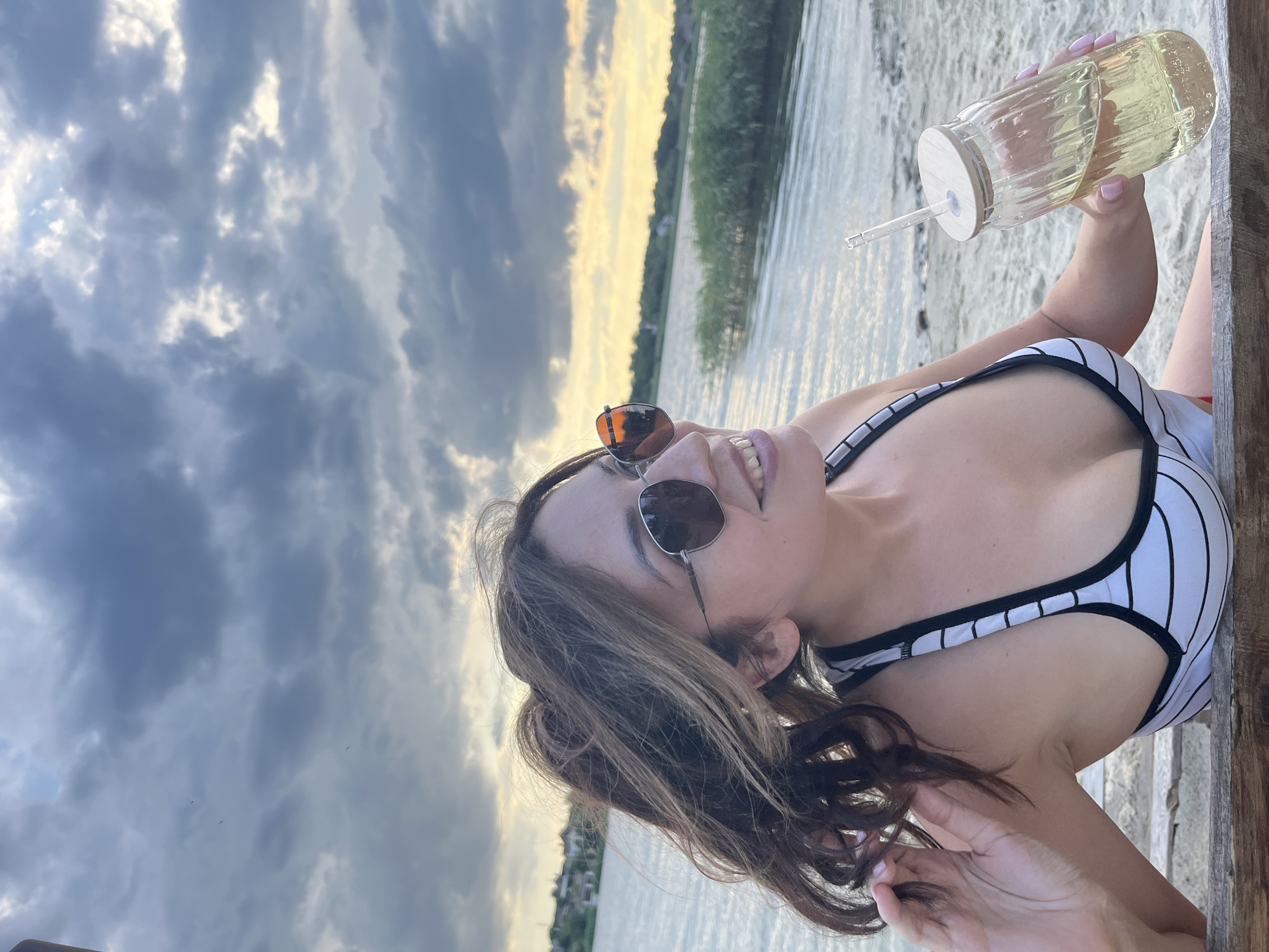A woman is sitting on a wooden bench by the water. She is wearing a black and white striped bikini top and sunglasses. She is holding a glass cup with a straw and a yellow drink. The sky is cloudy with some yellow light from the setting sun. The background shows a body of water with trees and buildings on the opposite shore. The location is Lozuvatka, Ukraine.