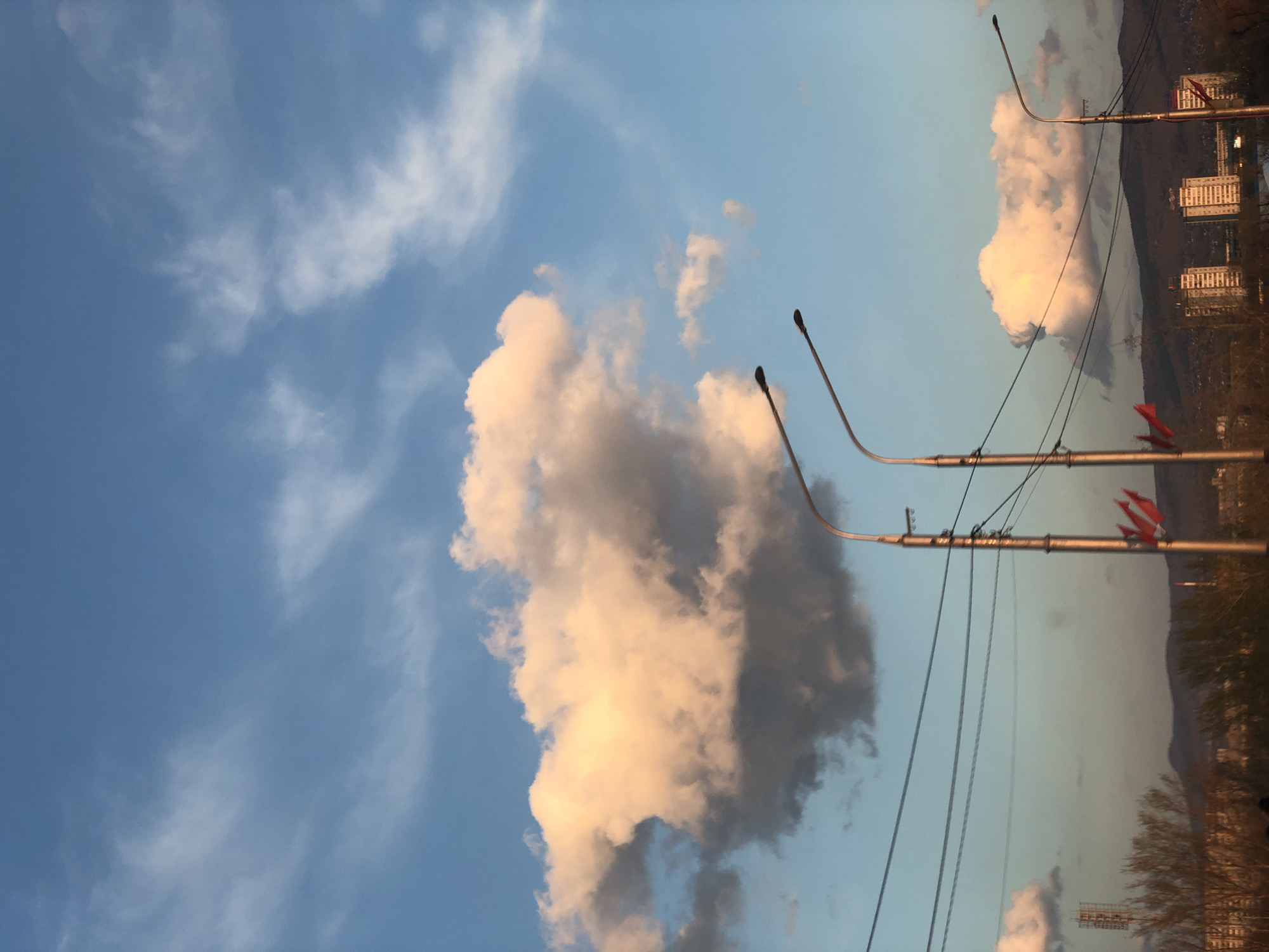 The image depicts a partially cloudy sky in Krasnoyarsk, Russia. In the foreground, streetlights are visible, as well as telephone poles. In the background, residential buildings are seen on the slope of a mountain. The time appears to be around sunset, given the color of the clouds.
