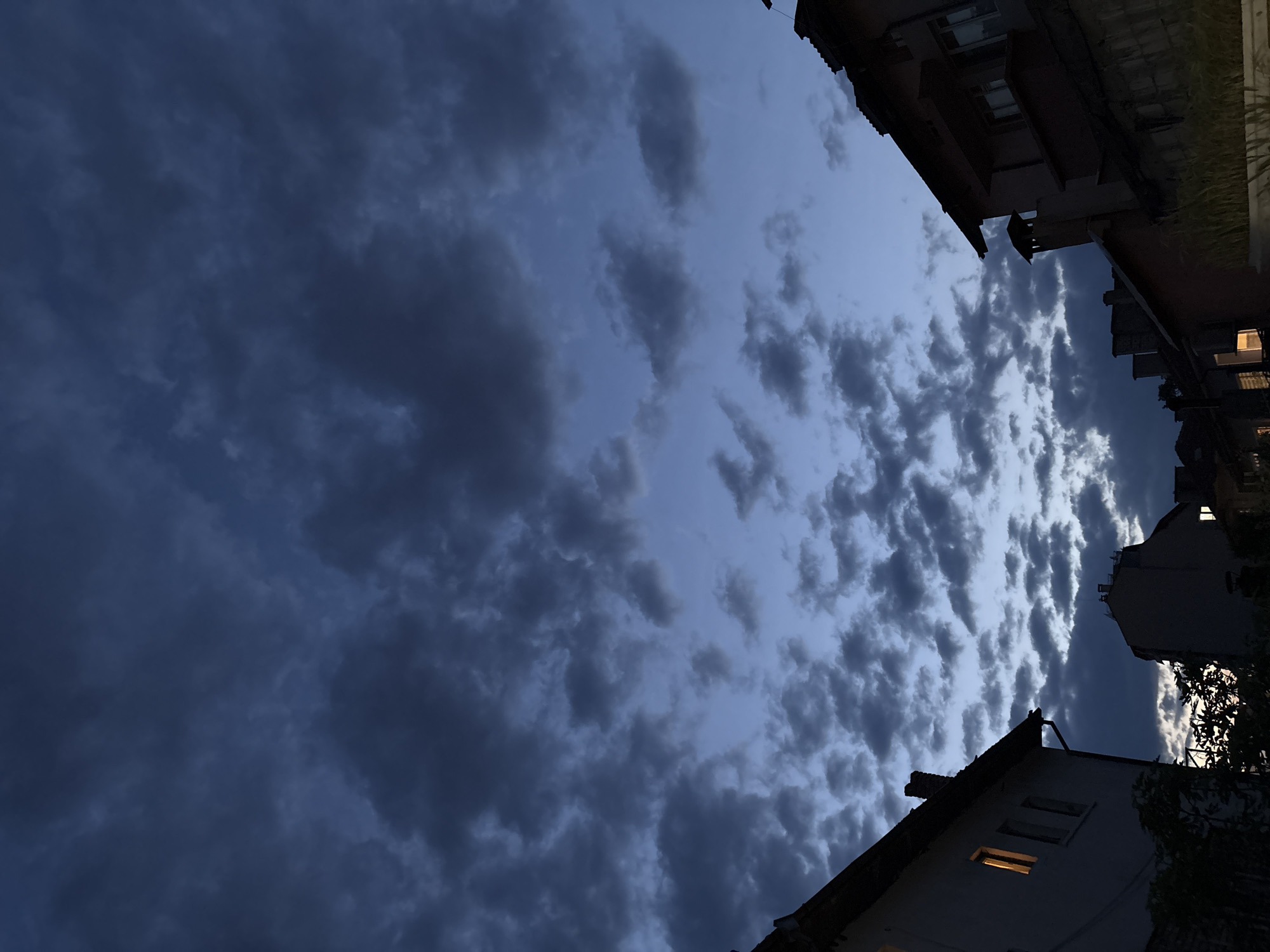 The image presents a low-angle view of a dusk sky above what appears to be a residential area in Varna, Bulgaria. Dominating the view is a dramatic cloudscape with varying shades of blue and grey, suggesting an approaching storm or the aftermath of a sunset. The clouds are a mix of textures, with some areas densely packed and dark, while others are lighter and more diffuse.
Framing the sky are the silhouettes of buildings, indicating an urban setting. A few windows are illuminated, suggesting activity inside. The architectural styles vary slightly, implying a mix of older and newer constructions typical of urban development. Foliage is visible around the buildings, adding a touch of greenery to the scene.
The overall composition suggests a moment captured in the transition from day to night, with the interplay of natural and artificial light creating a visually striking image.