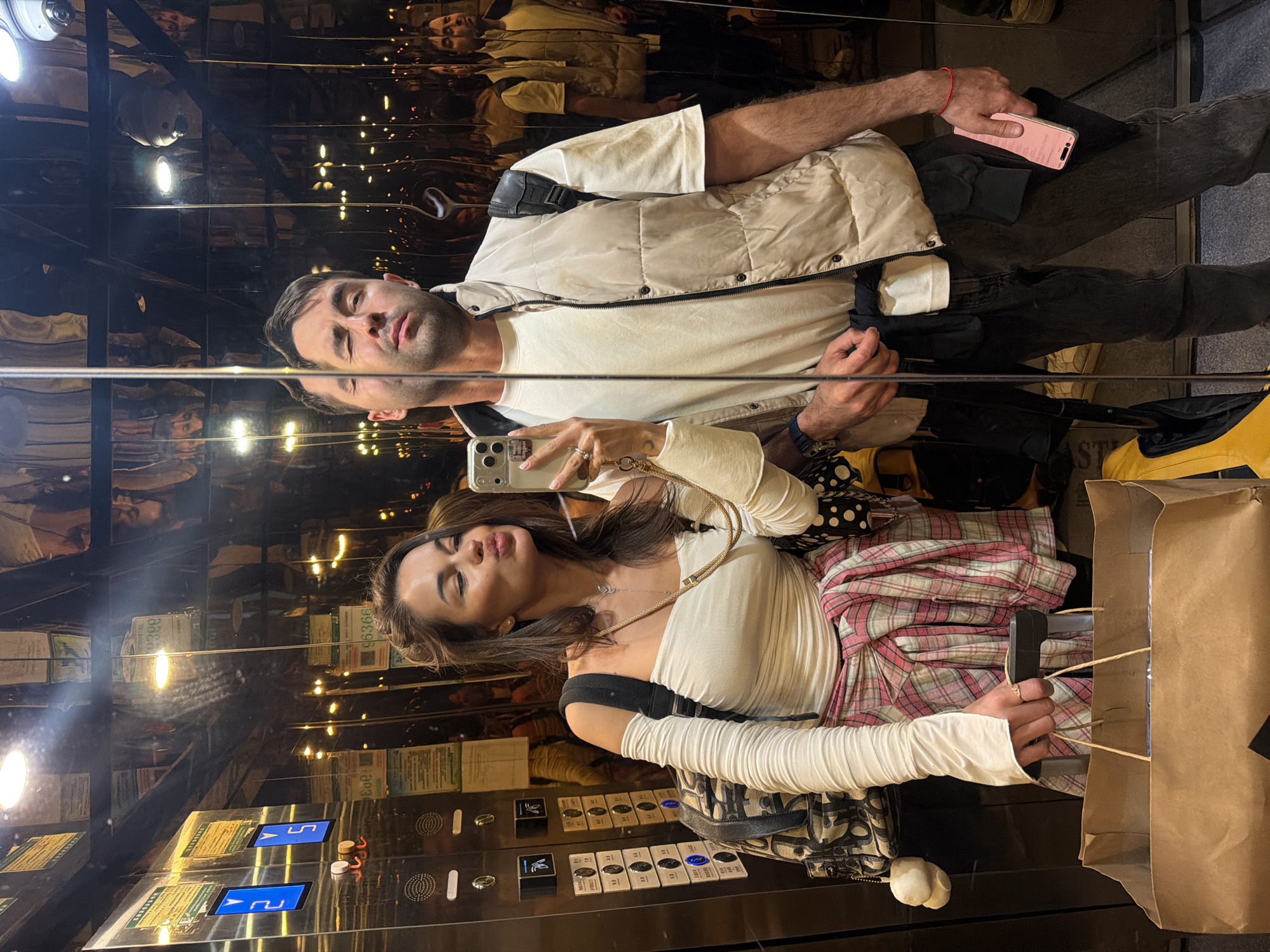 Two individuals are visible in an elevator with mirrored walls, capturing a selfie. The woman holds a smartphone in her right hand, her lips pursed for the photo. She wears a light-colored top, long sleeves, and a pink plaid skirt. A shopping bag is visible to her right. The man stands to her left, also visible in the mirrored reflection. He wears a light-colored shirt, a vest, and dark pants. His right hand holds a pink phone. The elevator's control panel is visible, and the reflection shows the city environment, possibly Zhangjiajie, China, due to the unusual elevator location and appearance. The light inside the elevator is dim, but the reflection reveals a brightly lit street scene.