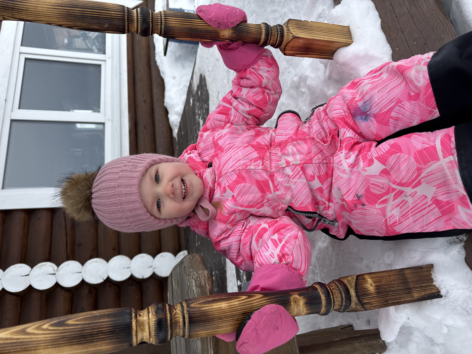 In Crokhovo, Russia, a young child is depicted outdoors in a snowy environment. The child, approximately two to four years old, has light skin, blue eyes, and light hair, and is wearing a light pink ribbed knitted hat with a large, dark brown fur-like pom-pom. A pink string from the hat is visible under the child's left ear. The child is dressed in a vibrant pink and white patterned snowsuit with black ankle cuffs and pink insulated mittens. The child is smiling broadly, facing the camera, with both hands grasping dark brown, turned wooden posts. These posts appear to be part of a larger wooden structure, possibly a swing or porch railing. The child is seated on a wooden surface partially covered by snow. In the background is a log cabin-style building with visible horizontal logs. A white-framed, double-paned window is present on the building's facade. To the left of the child, a vertical series of white, decorative, scalloped wooden elements is attached to the log wall. The ground is covered in snow. A faint blue smudge is visible on the right thigh area of the child's snowsuit.