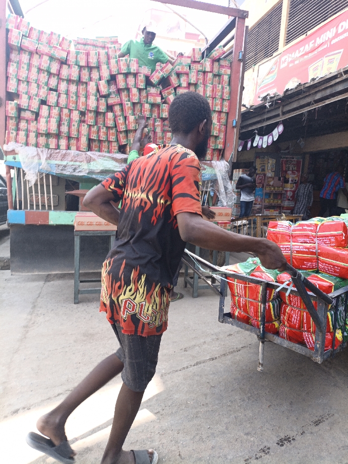 The image captures an active street-level commercial scene in Fagge C, Nigeria. In the foreground, a dark-skinned male, facing away from the viewer, is propelling a metal handcart. He wears a black t-shirt with red and yellow flame graphics, plaid shorts, and grey slide sandals. The handcart is loaded with multiple bundles wrapped in red material, secured with white strapping; text on the bundles partially reads "NEW HOP INTERNATIONAL TRADE".
In the midground, a large truck is parked, its bed filled with numerous stacked cardboard boxes featuring a distinct green and pink/red pattern. A dark-skinned male, wearing a green t-shirt and white cap, stands atop these boxes within the truck, actively handling them. Another individual's leg, also in green attire, is partially visible near the truck's rear.
The background features a storefront with a prominent red sign partially legible as "MAZAF MINI D...". The store's interior displays various packaged goods. Several individuals are discernible within or near the store's entrance. The ground is paved, showing tire marks and general wear. Shadows suggest outdoor daylight conditions.