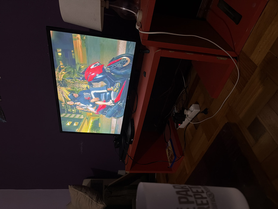 An indoor scene in Vila Real, Portugal, depicts a dimly lit residential room, illuminated primarily by a flat-screen LG television displaying a vibrant image. The television screen shows a man and a woman seated on a red scooter. The man, in a dark hoodie with white stripes and yellow-soled shoes, is at the front, with the woman, in a light-colored top, behind him. The background suggests an urban night setting with illuminated buildings and palm trees.
The television rests on a red, open-shelf entertainment unit. A black electronic device, possibly a media player, is situated on this unit below the screen. To the right, an identical red open-shelf unit supports a table lamp with a white fabric shade. To the far left, a portion of a light-colored cushion from a sofa or armchair is visible, adjacent to a vertical appliance. Numerous cables are visible, mostly tangled below and behind the red entertainment unit, connecting to a white power strip with several European-style plugs on the parquet wood floor. In the immediate foreground, a white mug with dark, partially visible lettering ("E PAR REPET") rests on a dark surface, likely a coffee table. The walls are painted a dark, possibly purple, color.