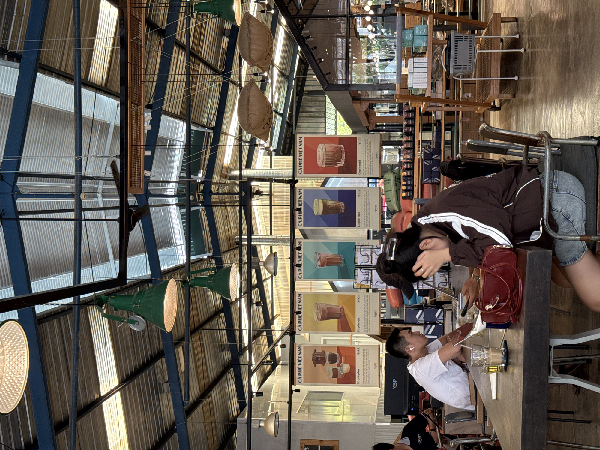 The interior of a Vietnamese café, likely in Da Lat, Vietnam, showcases a high ceiling constructed with corrugated metal, supported by blue steel beams. Hanging from the beams are several lamps, some enclosed in woven basket-like shades. A row of vintage-style coffee posters labeled "Cà Phê Việt Nam" (Vietnamese Coffee) is displayed along the wall, with illustrations of coffee drinks. A table occupies the foreground, where a young woman in a brown jacket and denim shorts is seated next to a red purse. She is leaning forward, resting her head in her hand. A young man in a white shirt is seated beside her, holding a straw and looking down towards a glass of iced coffee on the table. Shelving units stocked with products stand in the background. The overall atmosphere is casual and modern, with a focus on coffee culture.