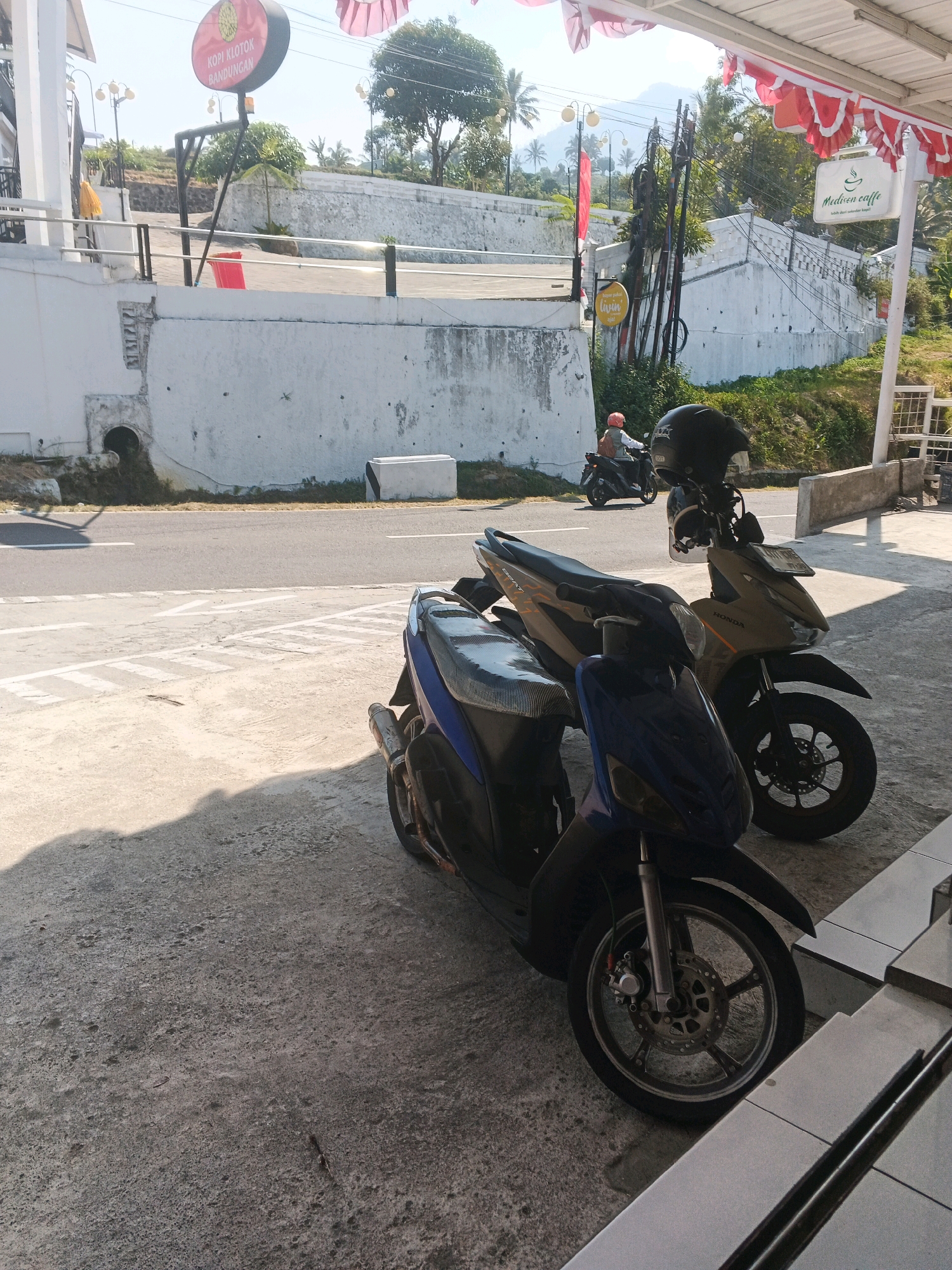 A motorcycle is parked on a sidewalk in front of a building. The motorcycle is blue and silver with a black seat. The building is white and has a red sign that says "KOPI KLOTOK BANDUNGAN". There is a person on a motorcycle in the background riding on the road. The sky is blue and there are some trees in the background. The image appears to have been taken in Pakopen, Indonesia.