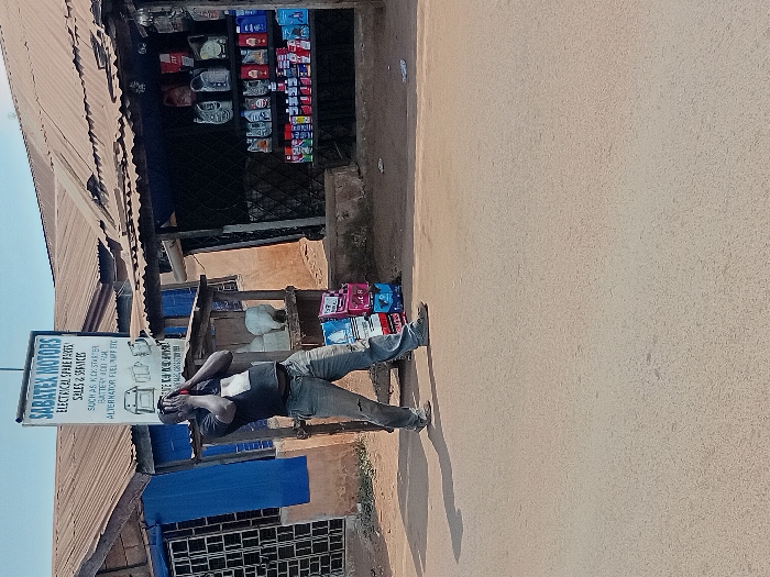 The image depicts a street scene in Ayangba, Nigeria, under clear daylight conditions. A single male subject, dark-complexioned, stands centrally on an unpaved road surface. He is wearing a dark t-shirt and light-colored trousers. His hands are raised, holding a red mobile phone to his right ear, thereby obscuring his face. A distinct shadow is cast to his left.
Behind the subject and to his left, a single-story building with a corrugated metal roof and blue window frames is visible. A prominent vertical sign affixed to a pole in front of this building reads "SABATEX MOTORS" at the top, followed by "ELECTRICAL SPARE PARTS SALES & SERVICES," with a list of items including "KICK STARTER," "BATTERY," and "FUEL PUMP."
To the subject's right, an adjacent single-story structure also with a corrugated metal roof functions as a retail establishment. Its open storefront displays multiple shelves stocked with various bottles of motor oil and other automotive fluid containers; "JTT" branding is visible on some products. Several stacked boxes, some appearing red or pink, are on the ground near the shop entrance, to the right of the subject. A white, indistinct object is positioned behind the subject, near the entrance of the right-hand shop.