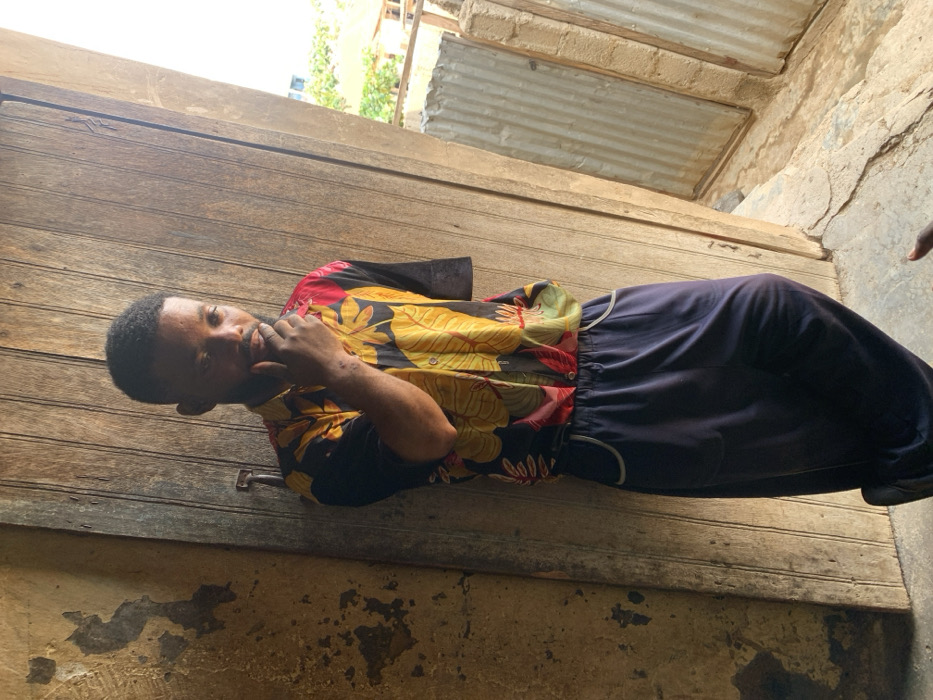 A young male, with a short beard and dark hair, stands in what appears to be an open doorway or alcove in Okene, Nigeria. He wears a short-sleeved button-up shirt with a black base and vibrant yellow, red, and green floral patterns, paired with dark blue wide-leg trousers featuring white piping around the pockets. His right hand is raised, with fingers touching his chin/mouth area. His left arm is not fully visible, appearing to terminate above the elbow. To his immediate left is a weathered wooden plank wall or door, secured with a visible metal handle. Further left, an adjacent wall shows significant peeling of light-colored plaster. To his right, beyond the doorway, vertical corrugated metal sheets are visible, partially obstructed by concrete blocks. Distant greenery and a light-colored building are discernible in the background. The ground beneath him is an uneven concrete or packed earth surface.