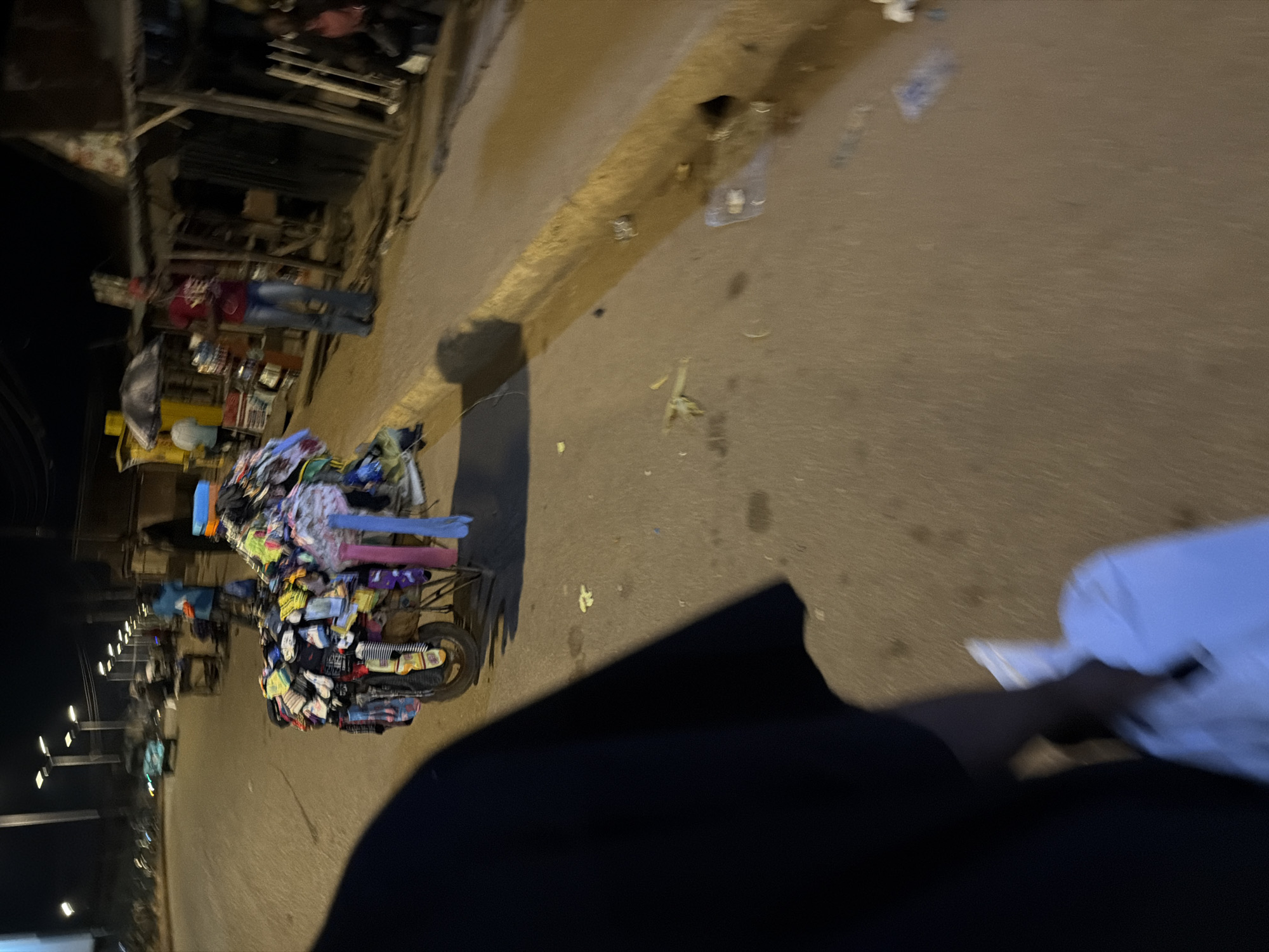 The image shows a nighttime street scene in Bukuru, Nigeria, with a focus on a mobile stall laden with clothing, primarily socks. The stall, mounted on a cart with a visible wheel, is positioned on the right side of a paved road. The clothing is a colorful assortment of socks, neatly folded and displayed.
In the foreground, the dark silhouette of a person is visible, carrying a white plastic bag. To the right of the stall, a person wearing a red t-shirt and blue jeans stands, appearing to be the vendor or an associate. Behind the stall, there are some simple structures, possibly market stalls or shops, constructed with corrugated metal and wood. The environment suggests a bustling marketplace or a busy street corner at night. Streetlights illuminate the scene, casting long shadows and creating a warm, yellow glow on the asphalt.
In the background, other people are present, some appearing to be shoppers or passersby. A vehicle, possibly a car, is dimly visible in the distance, and other streetlights line the road. The overall impression is of a lively urban environment at night, with commercial activity taking place. There is some debris scattered on the road and sidewalk, typical of a public space. The focus on the clothing stall suggests it is a key element of the scene, highlighting local commerce and street vending.