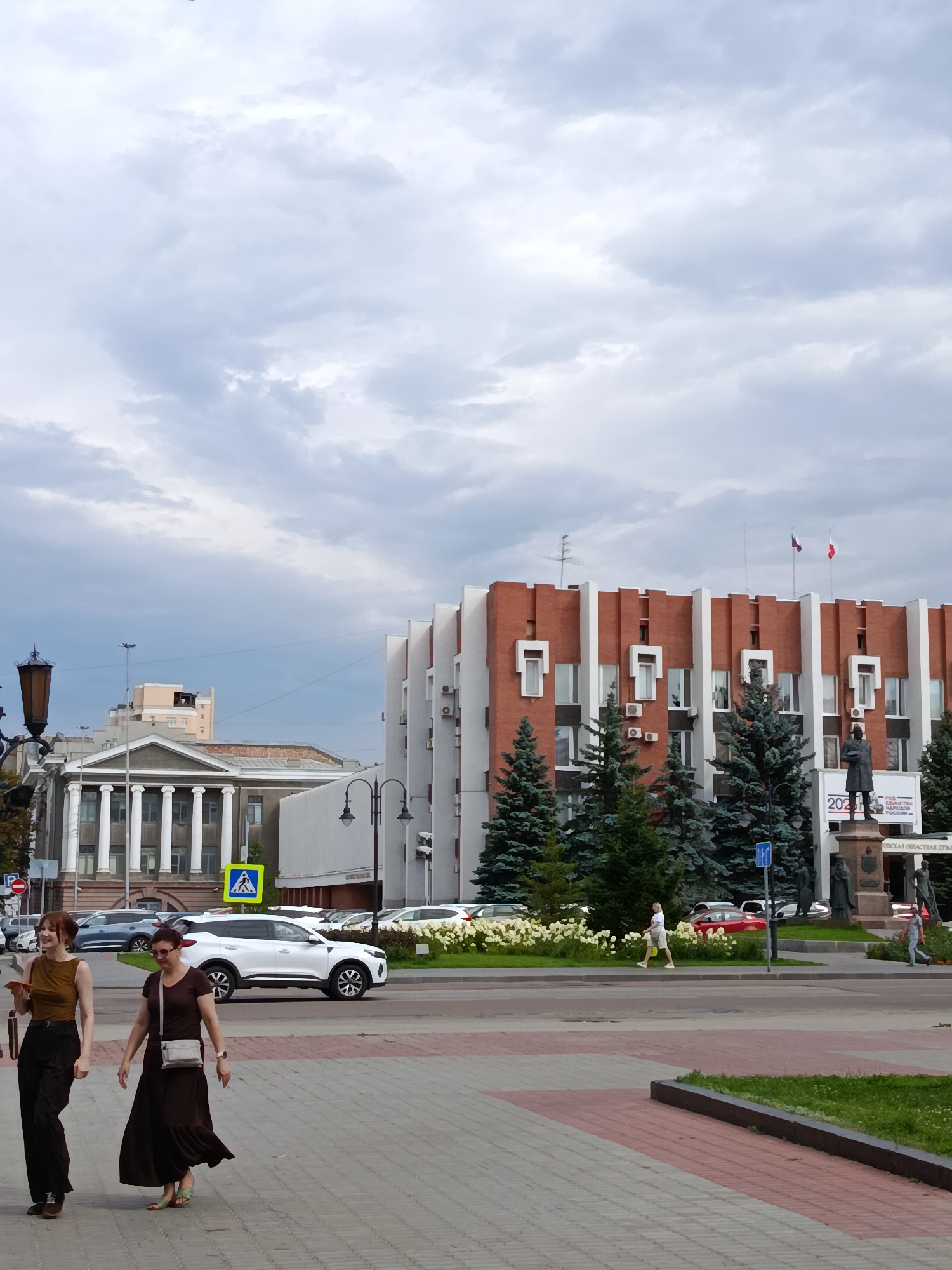 In Saratov, Russia, on a cloudy day, a street scene unfolds in front of a modern brick building with white trim and a statue. Two women walk across a patterned sidewalk in the foreground. The woman on the left wears a mustard-colored top and dark pants. The woman on the right wears a long brown dress. In the middle ground, a white SUV is parked, with other cars visible behind it. A pedestrian crossing sign is posted on a yellow background. Trees and flower beds line the street. The brick building features a repeating pattern of white rectangular elements and several windows. A statue stands in front of the building, accompanied by a banner that reads "2023 YEAR OF UNITY OF THE PEOPLES OF RUSSIA". Another building with classical columns is visible to the left. The sky is overcast with gray clouds.