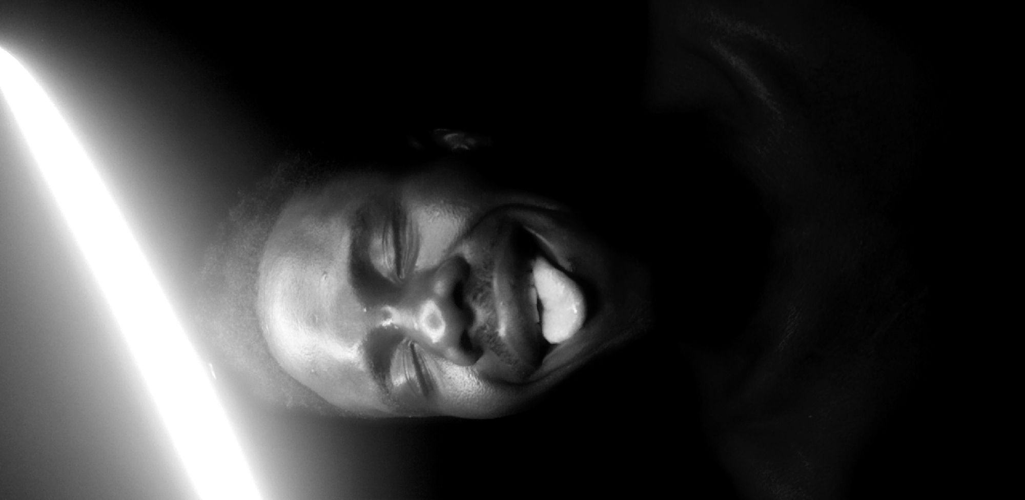 A black and white, close-up photograph depicts a person's face illuminated by a bright light on the left side of the frame. The person's eyes are closed, and their mouth is open, revealing their tongue. The background is dark and indistinct. The image appears to be taken indoors with artificial lighting. The location of the photo is unknown and can not be determined from the image. The city of Narayi, Nigeria, is also unknown.