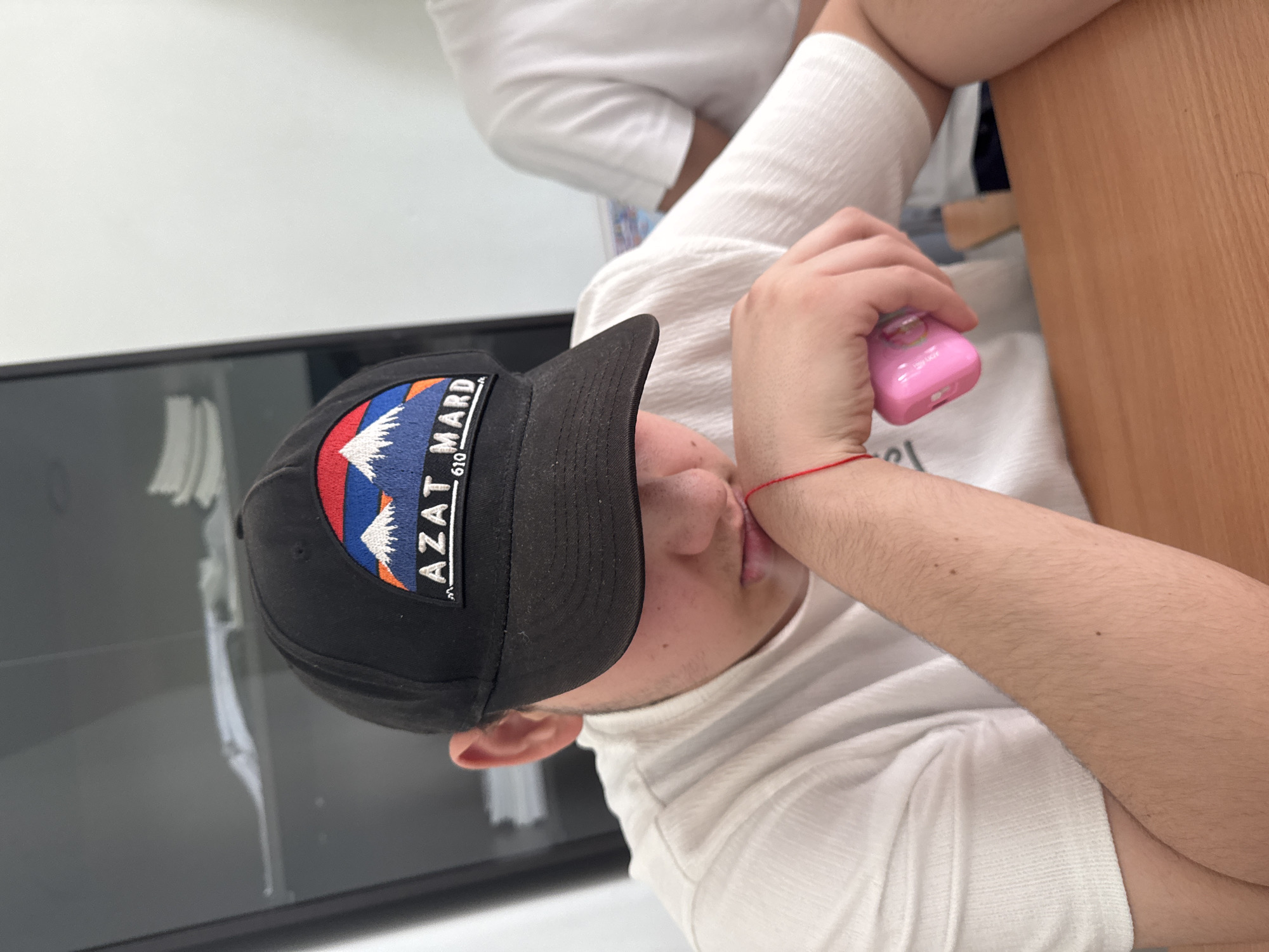 An overhead, vertically oriented shot captures a person, likely a young adult male, in an indoor setting. The individual is wearing a black baseball cap with a prominent embroidered patch and a white long-sleeved, ribbed shirt. They are positioned next to a light-colored wooden surface, possibly a desk or table, which occupies the right side of the frame.
The person's head is tilted, and their left arm is bent, holding a small, bright pink electronic device. A thin red string or bracelet is visible on their left wrist. Their face is partially obscured by the cap's brim and their arm, but their ear and part of their cheek are visible. Their mouth is slightly open, with a hint of their tongue protruding.
In the background, a white wall is visible on the left, and a dark, reflective surface, possibly a window or large monitor, runs along the bottom left. The embroidered patch on the black cap features a multi-colored mountain landscape design. Text on the patch clearly reads "AZAT MARD" and below it, partially visible, "610". "Azat Mard" translates to "Free Man" in Armenian. The pink device held in their hand shows a faint graphic, resembling a strawberry or similar fruit, and possibly some small text like "Lovely Cute". The person's arm exhibits light hair and a couple of small moles. The scene suggests a casual moment indoors in Yerevan, Armenia.