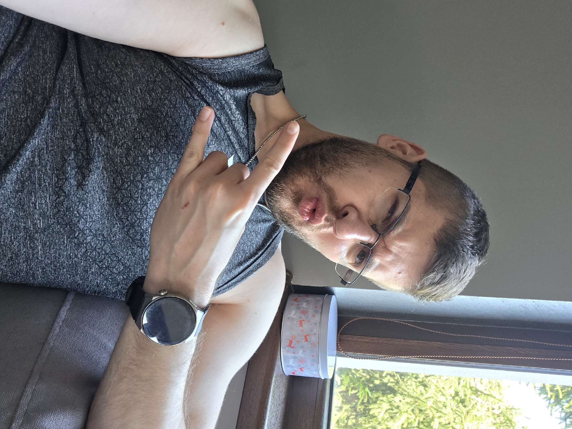 A man is posing indoors with his head turned to the right and slightly tilted. He wears glasses, a gray t-shirt, and a watch on his left wrist. His left hand is raised in a "rock on" hand gesture. He is making a playful face, sticking his tongue out slightly. A decorative tin can sits on a windowsill beside him, and a window with a view of greenery is visible behind the tin. The location is not discernible as being in Yagodovo, Bulgaria, as there are no identifying landmarks or contextual clues within the image.