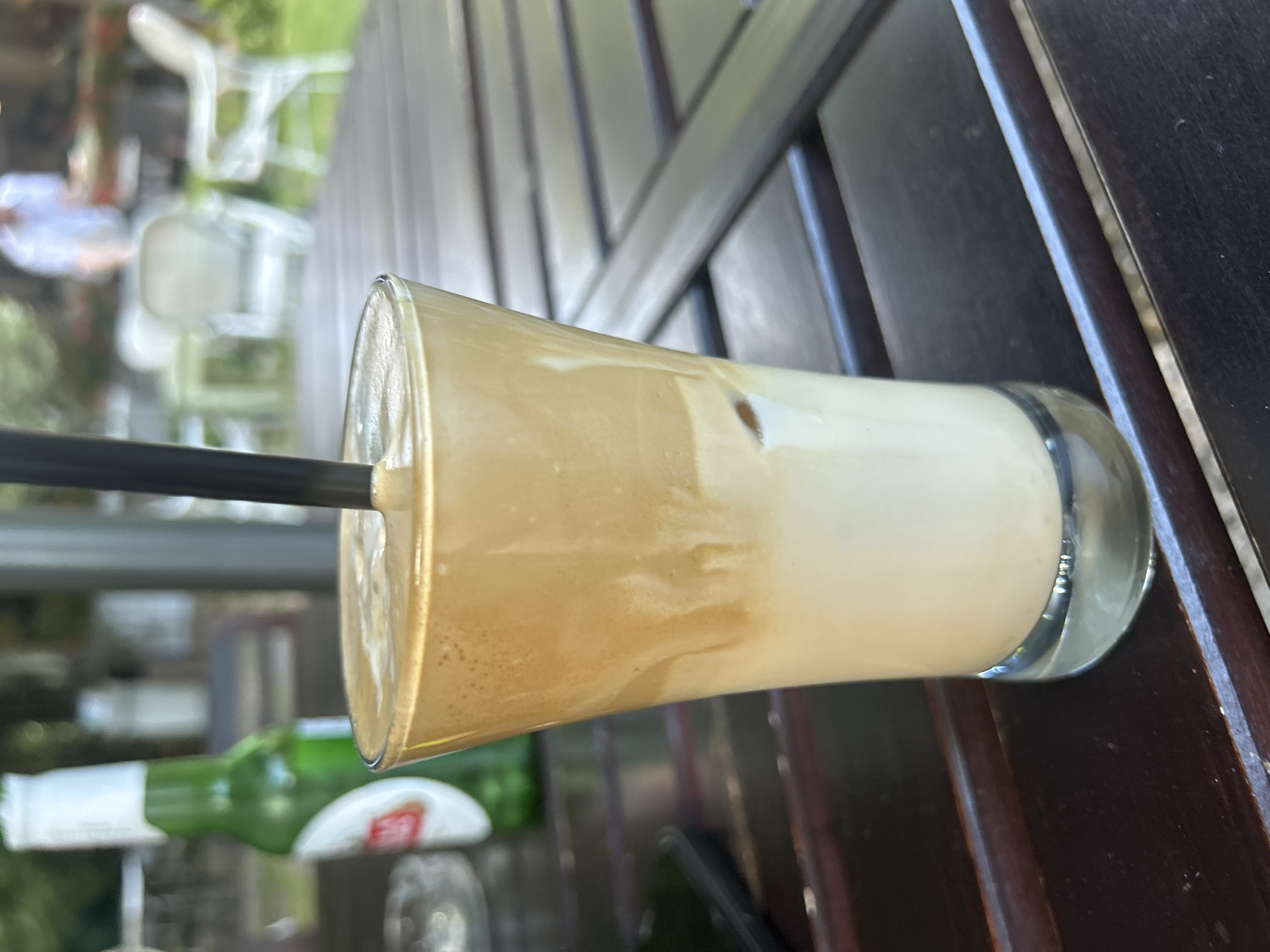 The image shows a glass of Dalgona coffee with a black straw, placed on a dark wooden table. In the background, out of focus, is a green bottle of beer with a white label, and a blurred outdoor setting with tables, chairs, and greenery, suggesting a cafe or restaurant. The cafe appears to be in the city of Varna, Bulgaria, a popular seaside resort known for its outdoor dining establishments.