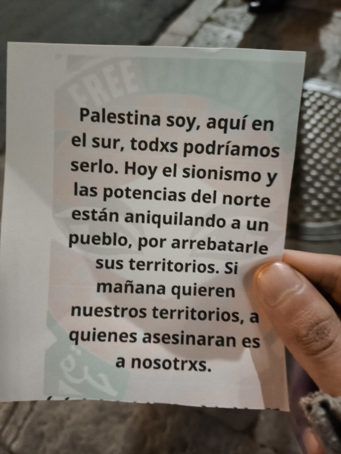 A close-up photograph displays a white paper flyer, held upright in the lower right by a human hand, specifically the thumb and index finger, with light to medium brown skin tone. The flyer features prominent black text in Spanish, stating: "Palestina soy, aquí en el sur, todxs podríamos serlo. Hoy el sionismo y las potencias del norte están aniquilando a un pueblo, por arrebatarle sus territorios. Si mañana quieren nuestros territorios, a quienes asesinaran es a nosotrxs." This translates to: "I am Palestine, here in the south, we could all be. Today Zionism and the northern powers are annihilating a people, to snatch their territories. If tomorrow they want our territories, it is us they will assassinate." Faint green and orange graphic elements, along with barely visible "FREE" and Arabic script, form a background on the flyer, suggesting a "Free Palestine" message. The background behind the hand and flyer is out of focus, depicting a dark, textured urban environment, possibly a street or sidewalk, with dim ambient lighting. A partial view of a dark grey garment is visible near the holding hand. No specific landmarks or unique features are present to determine a precise location within the city of La Paz, Bolivia.