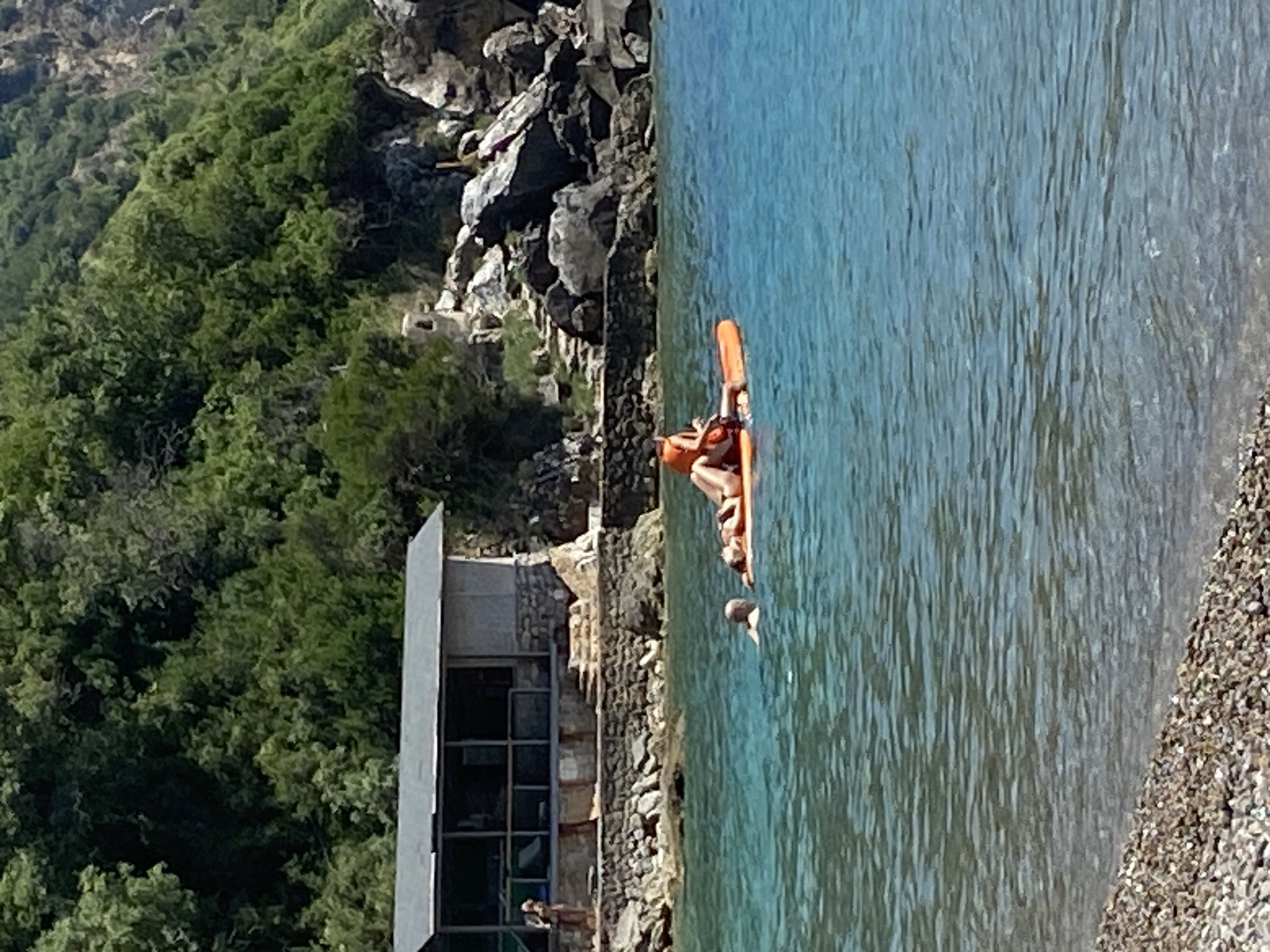 In the city of Prijevor, Montenegro, on a sunny day, two people are on an orange inflatable mattress in clear turquoise water. One person, wearing an orange life vest, is lying down, while another person is partially submerged in the water next to the mattress. In the background, a small building with a gray roof and stone walls is situated near the rocky shoreline, which is backed by a dense green forest on a hillside. The water is calm, with small ripples on the surface, and the shore is covered in pebbles.