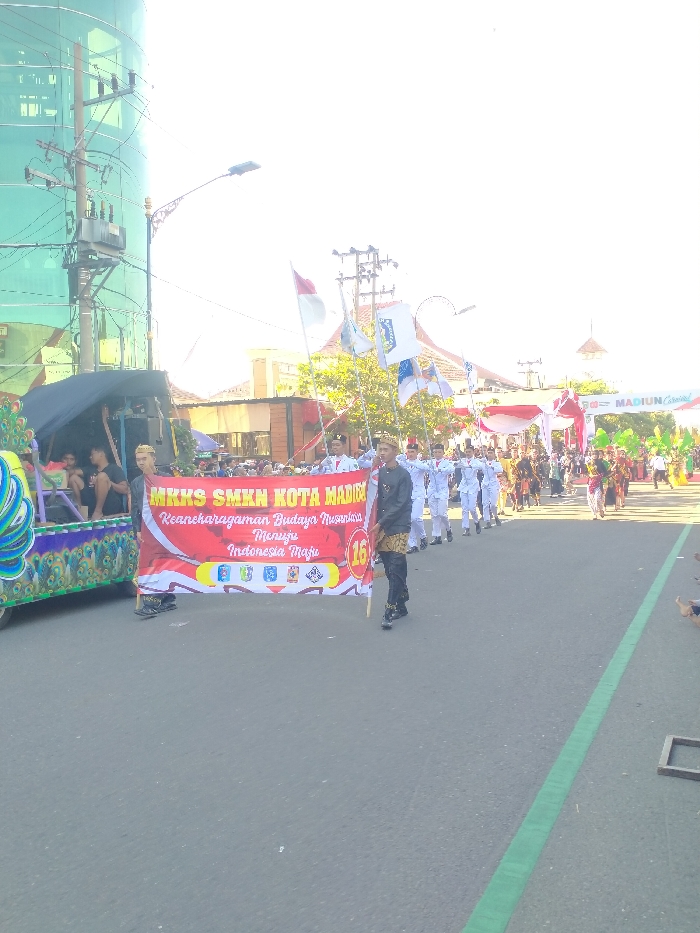 An outdoor parade scene unfolds on a paved street in Madiun, Indonesia. In the foreground, a dark gray asphalt road occupies the lower third of the frame, featuring a distinct green line extending longitudinally on the right side. A red and white banner, held horizontally across the road by two individuals, is central. The banner displays yellow text "MKKS SMKN KOTA MADIUN" and white text "Keanekaragaman Budaya Nusantara Menuju Indonesia Maju" (Diversity of Nusantara Culture Towards a Developed Indonesia), along with a red circle containing the number "16" and four small icons.
The individual holding the banner on the right wears a dark jacket with gold accents, dark pants, and a dark cap. To the left of the banner, a decorative float, primarily green and blue with peacock-eye patterns, moves along, carrying at least two partially visible individuals.
Behind the banner, a uniformed group marches in formation. This group, consisting of approximately 10-15 individuals, wears white long-sleeved uniforms with red detailing and white headwear. Several members carry poles with flags, including the red and white flag of Indonesia and multiple blue and white flags with a distinct crest. Following this group, a longer procession of individuals dressed in various traditional Indonesian attire proceeds down the street.
In the background on the left, a tall building with a curved, reflective green glass facade is prominent. A utility pole with multiple cross arms, transformers, and a network of overhead wires stands in front of it. A decorative street lamp extends over the road from the left. On the right background, buildings with orange-tiled roofs and green trees are visible, leading to a festive archway spanning the road. This archway is decorated with red and white fabric and green elements, displaying the text "MADIUN Carnival." The sky is intensely bright and overexposed, indicating strong overhead sunlight.