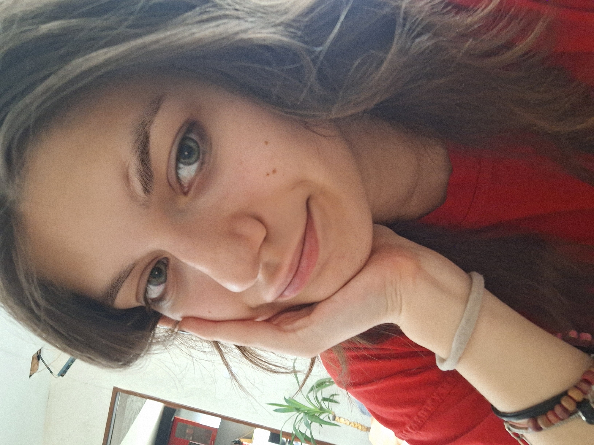 A woman with long brown hair and fair skin is resting her face on her left hand. She has green eyes and a slight smile. She is wearing a red shirt and several bracelets on her left wrist. The background shows a room with a mirror reflecting a red object, a green plant, and part of a white wall. The image is oriented at an angle. The location in Panagyurishte, Bulgaria is not evident from the image.