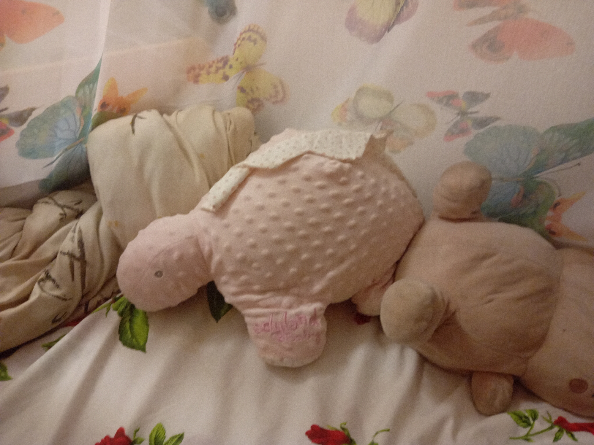 The image shows two stuffed animals, a pink turtle and a beige bear, lying on a white floral-patterned sheet. The turtle is in the foreground, with its head turned to the left. It has a textured pink shell and a white polka-dotted bib. The bear is to the right of the turtle, lying on its side. The background is a sheer curtain with a butterfly print. The scene appears to be indoors, in a bedroom or nursery. There is no indication of the location within the city of Chuhuiv, Ukraine.