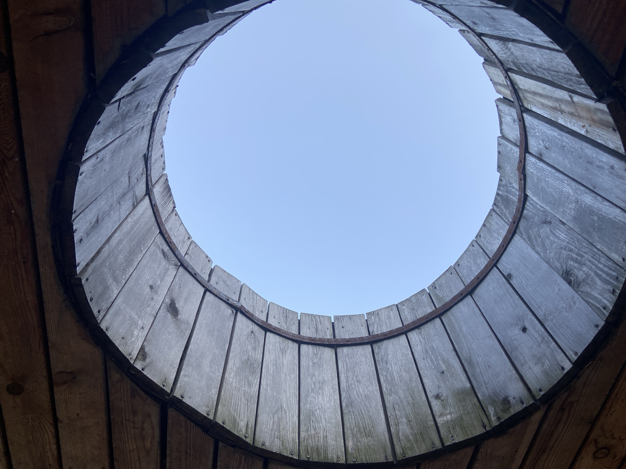 The image is an upward-looking shot through a circular opening formed by wooden planks and a metal band. The wooden planks are weathered and gray, arranged radially around the opening. The metal band appears rusted and is secured to the planks with small fasteners. Beyond the circular opening, a clear, pale blue sky is visible. The surrounding structure is composed of darker wooden planks. The scene is likely an outdoor setting, possibly a well, a manhole cover, or part of an architectural feature. No specific landmarks or identifying features of Balashikha, Russia are apparent in the image itself.