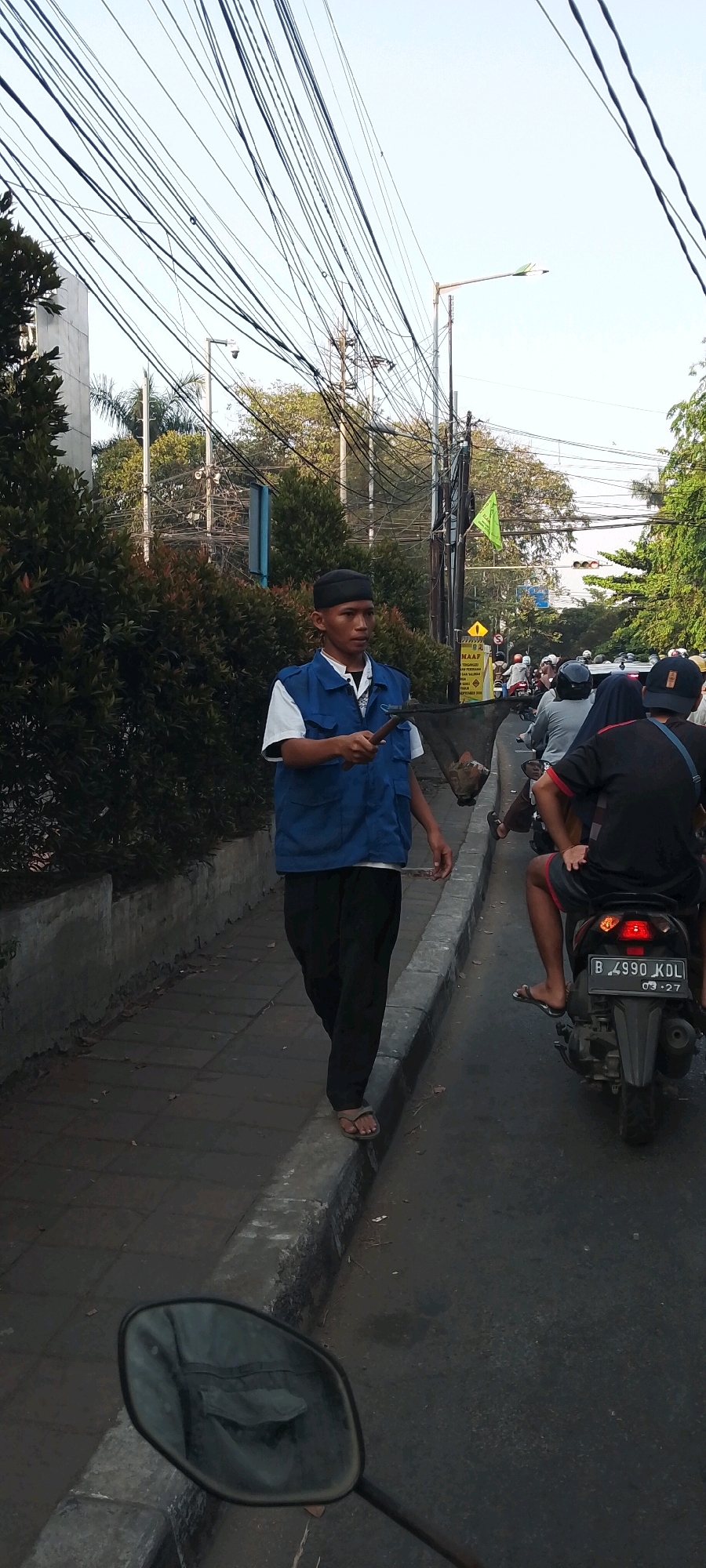 In Bekasi, Indonesia, a man wearing a blue vest, white shirt, and black pants walks on the sidewalk next to a busy road. He holds a tool with a net attachment. Vehicles, including motorcycles, are present on the road, indicating traffic. Overhead, numerous power lines crisscross the sky, and a street light is visible. The environment appears to be urban, with buildings and trees lining the street.
