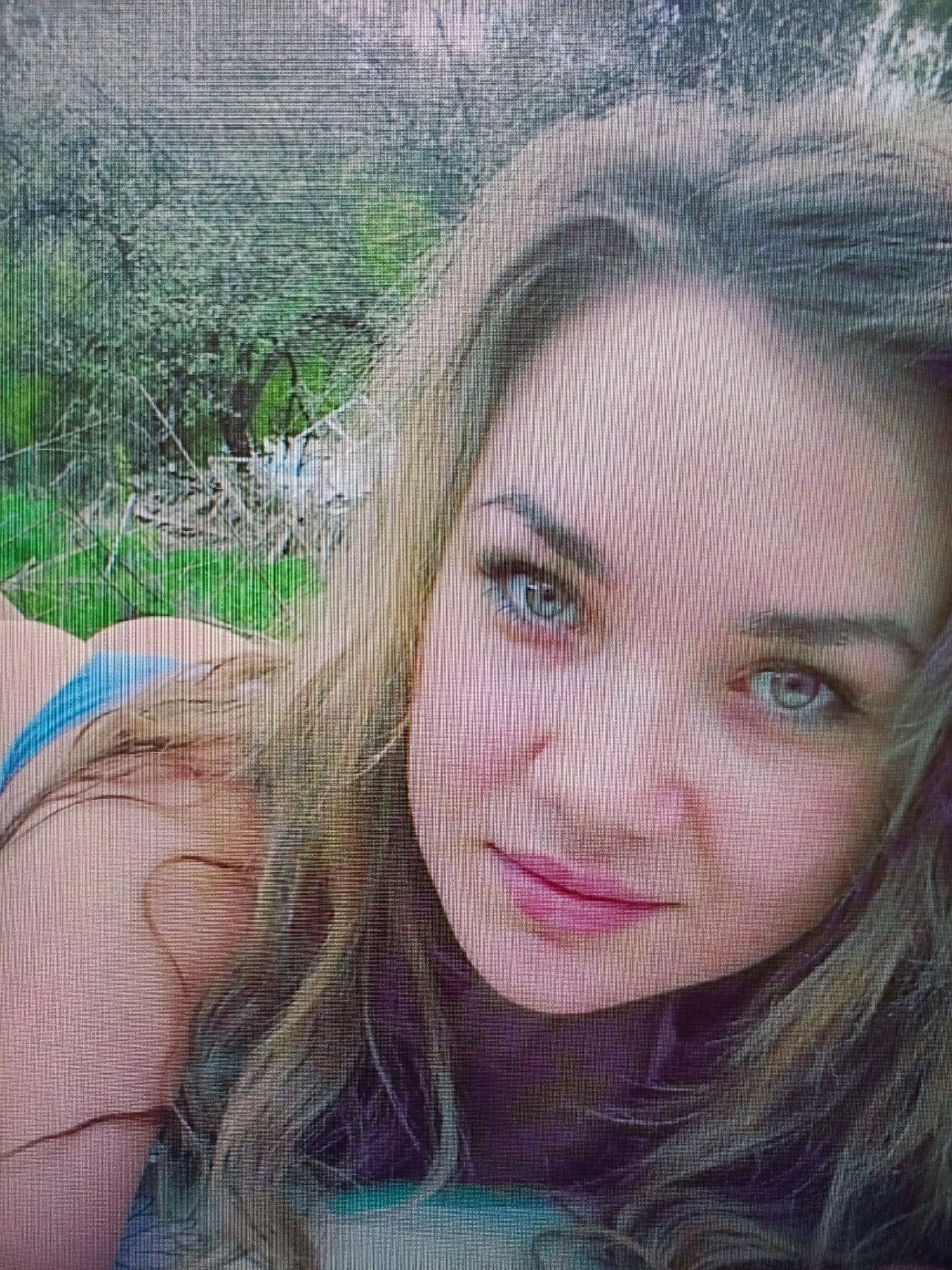 A close-up, low-angle selfie of a young woman with blonde hair and blue eyes lying on the ground. She is wearing a blue bikini top. The background appears to be a natural setting with greenery and some debris. The image resolution is poor, with visible pixelation and scan lines. The location is not specified in the image or its context.