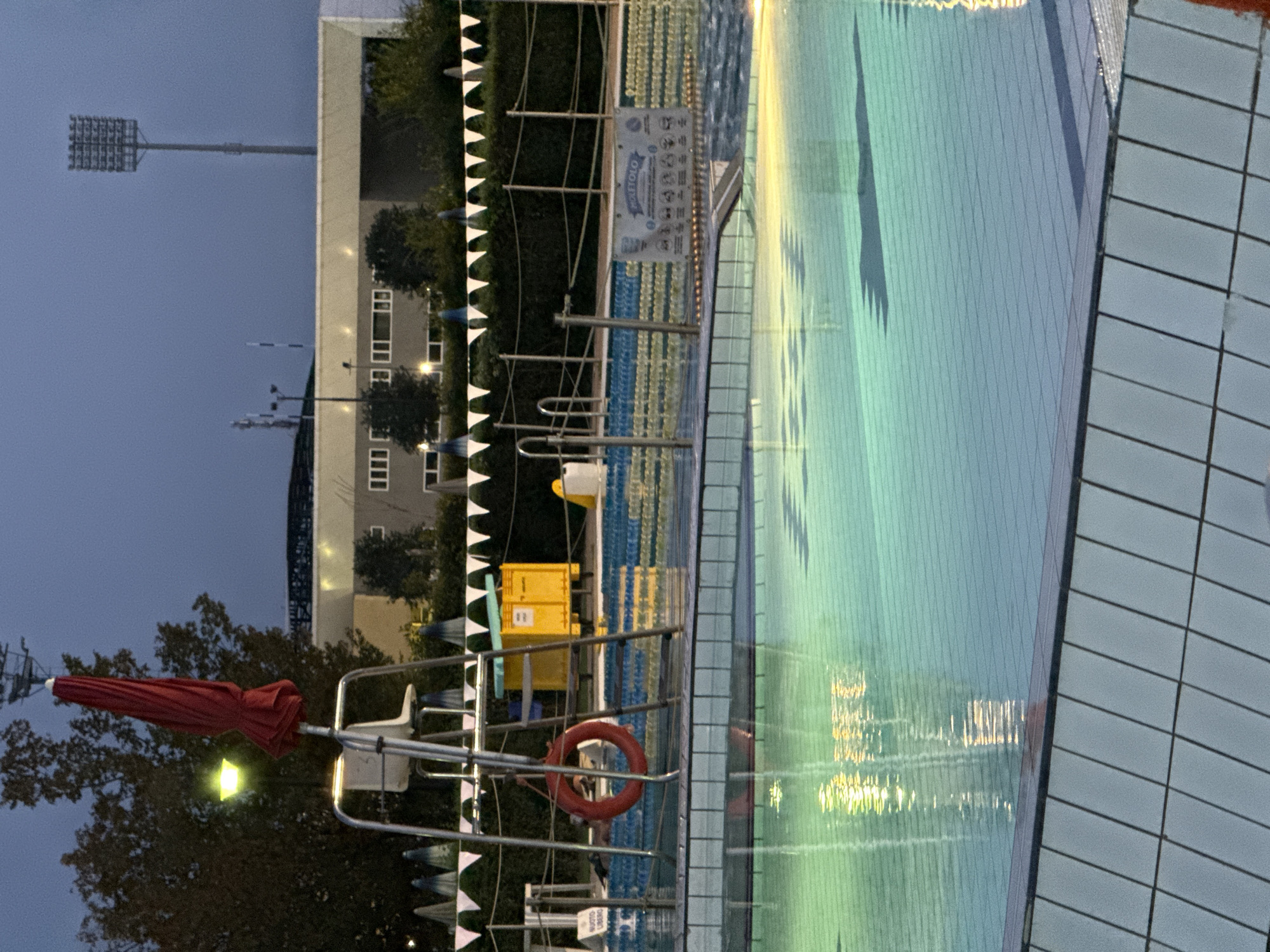 The image depicts an outdoor swimming pool area at dusk, possibly in Parma, Italy, as suggested by the presence of Italian text on a sign. In the foreground, the pool water is illuminated by underwater lights, casting greenish and yellowish reflections on the surface. The pool is bordered by a tiled edge. Behind the pool, a lifeguard chair with a red umbrella is visible. A red life preserver ring hangs from the chair. To the right of the lifeguard chair is a yellow box and a portion of a swimming lane marker. In the background, a building with multiple windows and a stadium light tower are present. The sky is a dusky blue. A banner with Italian text and pictograms is affixed to a railing near the pool's edge. Several triangular flags are strung along the perimeter of the pool area. The scene appears to be deserted.
