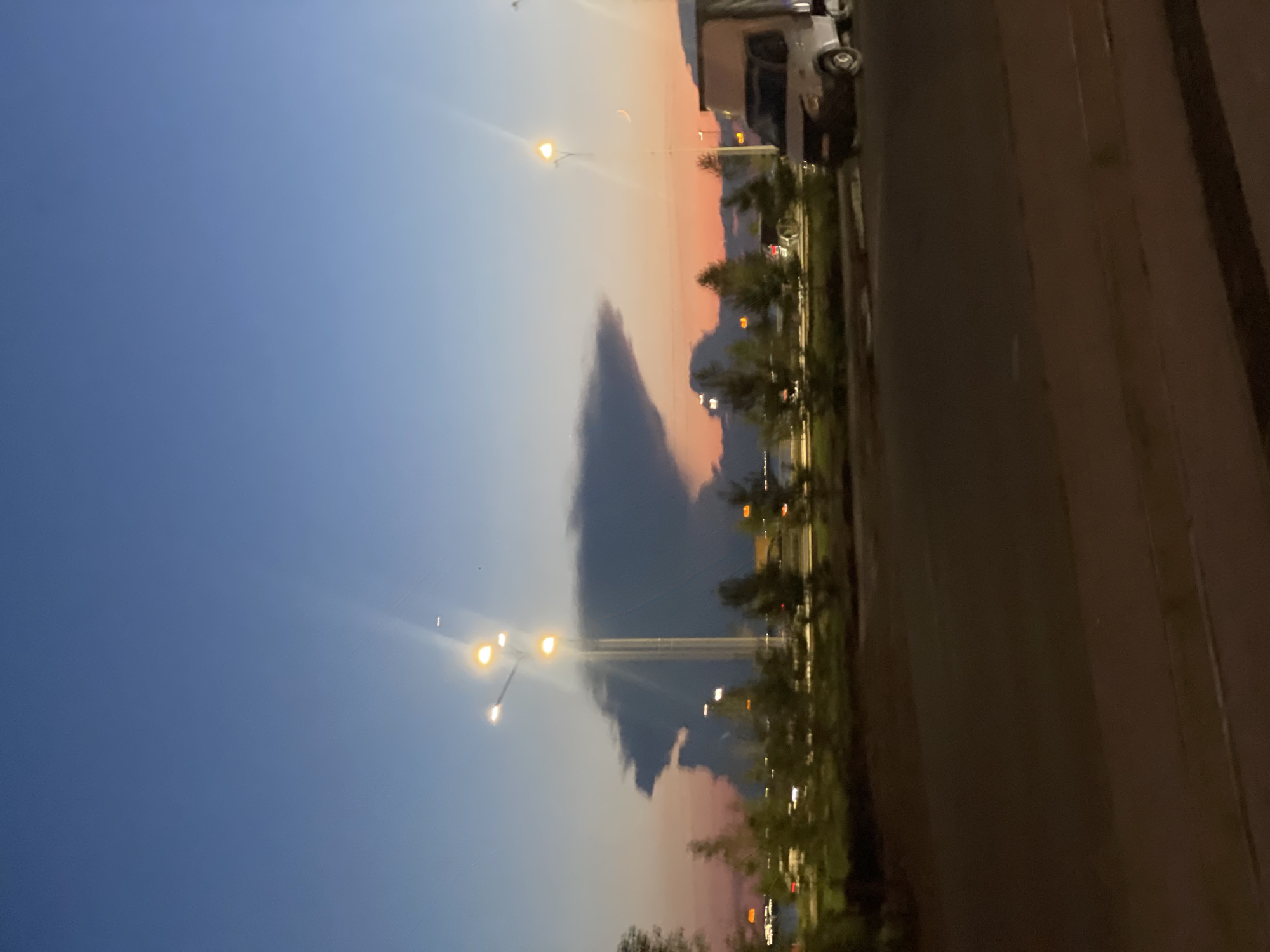 The image depicts a twilight scene in Kokshetau, Kazakhstan. A distinctive cloud formation, resembling a human profile with a prominent nose and chin, dominates the sky. In the foreground, a road is visible with a moving van and a streetlight with multiple illuminated bulbs. A line of trees and streetlights runs along the roadside, and in the distance, lights from buildings or a cityscape can be seen. The sky transitions from a deep blue at the top to shades of orange and pink near the horizon, indicating the setting or rising sun. The overall impression is a serene urban landscape at dusk, punctuated by the unusual cloud formation.