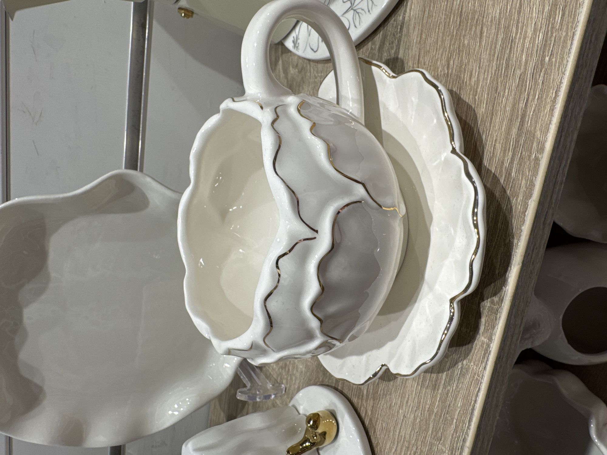 A close-up shot displays an elegant collection of white ceramic tableware, prominently featuring a teacup and saucer. The main teacup, made of gleaming white ceramic, has a distinct handle gracefully curved like a swan's neck. Its body is decorated with delicate, wavy gold lines that cascade down its sides, complementing the gold rim. The matching saucer, positioned behind the cup, boasts a ruffled or scalloped edge also outlined in gold.
The teacup is displayed upside down, hanging on a small, clear plastic stand, suggesting it is part of a retail or exhibition arrangement. To the left, a larger, plain white ceramic dish with a similarly ruffled edge is partially visible. The items are set against a backdrop of a light-colored wooden shelf with a visible grain, and a metallic vertical element is seen in the far background. Below the main display, another small white ceramic item with a golden, swan-like handle is partially visible, hinting at more items in the collection. The setting appears to be an indoor environment, likely a homeware store or a display area, characterized by artificial lighting. There are no people, visible text, or discernible time of day. The overall impression is one of sophisticated craftsmanship and decorative appeal.