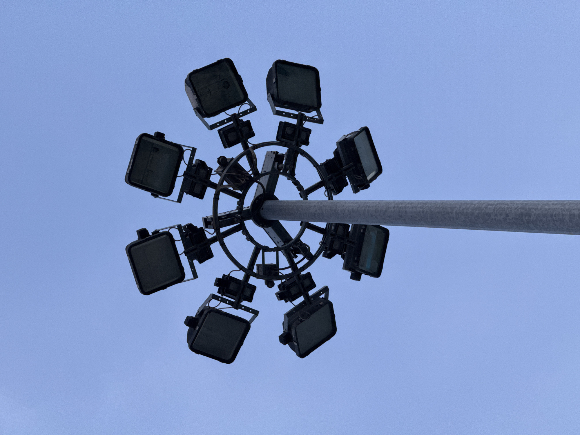 An outdoor, upward-facing, medium shot captures a lamppost topped with a circular array of spotlights against a clear, pale blue sky. The lamppost is a slender, grey cylindrical pole. Radiating from the top of the pole is a circular metal frame from which eight large, square spotlights are mounted at regular intervals. Each spotlight has a dark housing and a rectangular lens. The spotlights are angled in various directions, some pointing directly upwards, others outwards. The overall impression is of a powerful lighting fixture, likely for illuminating a large area such as a stadium or public square. The image does not provide any specific indicators of its location in Parma, Italy, or any surrounding urban context. There are no people, animals, or vehicles visible in the frame. The lighting suggests it might be dusk or dawn, or a heavily overcast day, as the sky is uniformly bright without any visible sun.