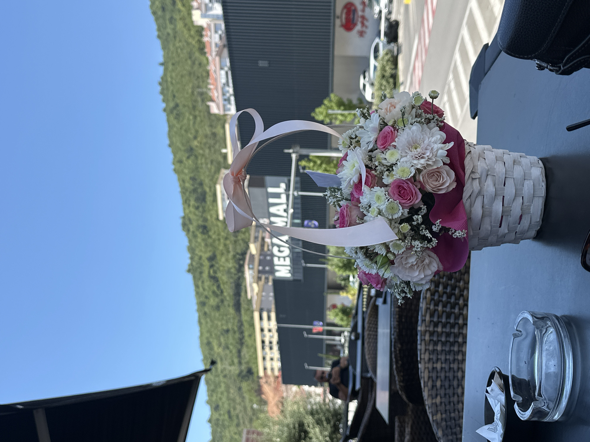 An outdoor cafe scene is depicted on a bright, sunny day with a clear blue sky. In the foreground, a table holds a clear glass ashtray filled with cigarette butts and paper wrappers, a small black bowl with more wrappers, and a white woven basket containing a bouquet of pink and white flowers adorned with a pale pink ribbon. The table is dark, and the chairs are made of wicker. In the mid-ground, a man wearing a dark t-shirt and sunglasses is seated at another table, looking at his phone. Behind him is a large building with the words "MEGA MALL" prominently displayed in white letters against a dark facade. Further in the background, a green, treed hill rises, dotted with buildings and their red tiled roofs. The scene appears to be located in Budva, Montenegro, likely near the Mega Mall, a shopping center situated at the base of the hills. Cars are visible on a road in front of the mall.