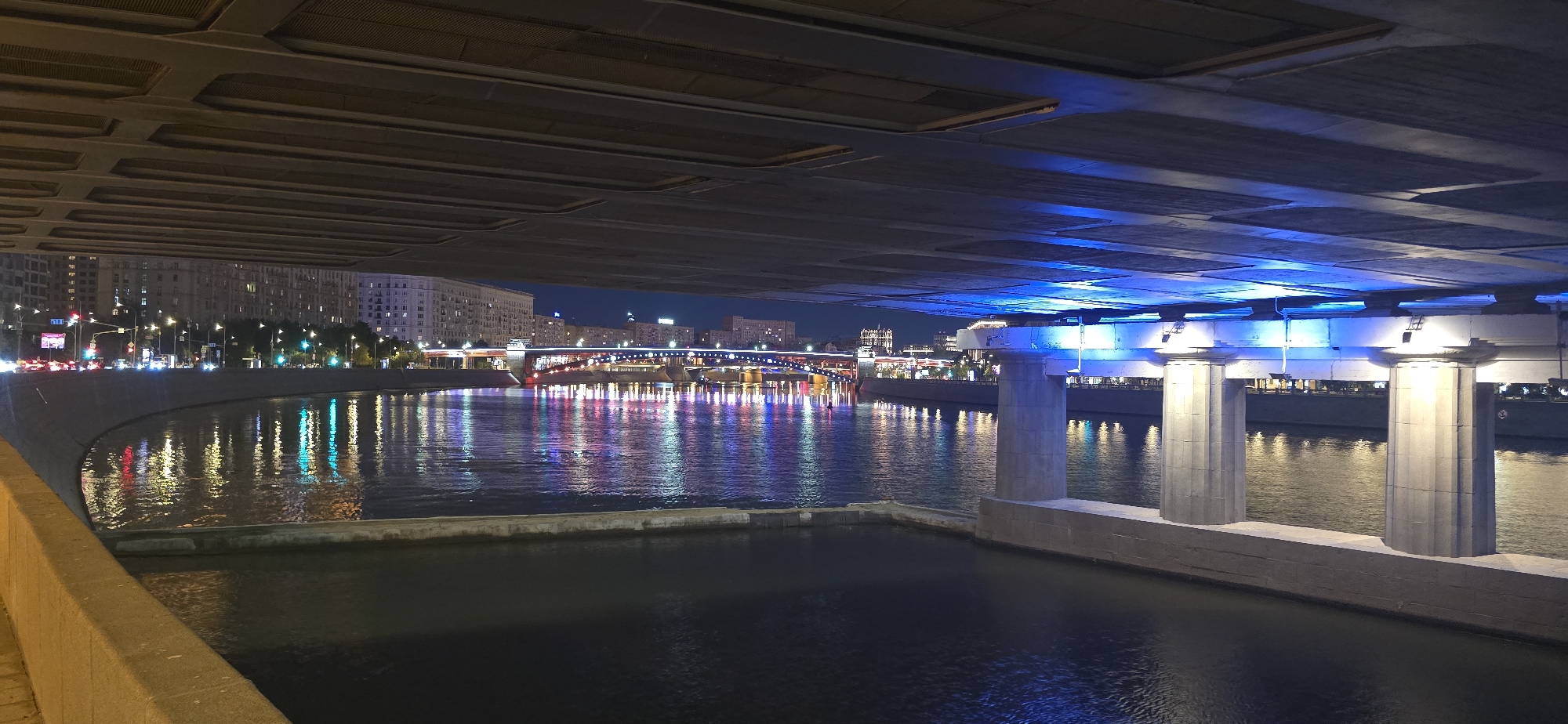 The image shows a view of the Moscow River at night, from beneath an overpass. The overpass is illuminated with blue lights, and its concrete structure is visible overhead. The river reflects the lights of the city and a bridge in the distance, which is decorated with colorful lights. On the left bank of the river, buildings are illuminated, and traffic lights and streetlights can be seen. On the right bank, there are also buildings and lights. There are no people visible in the image. The scene is static and captures a nighttime urban landscape.