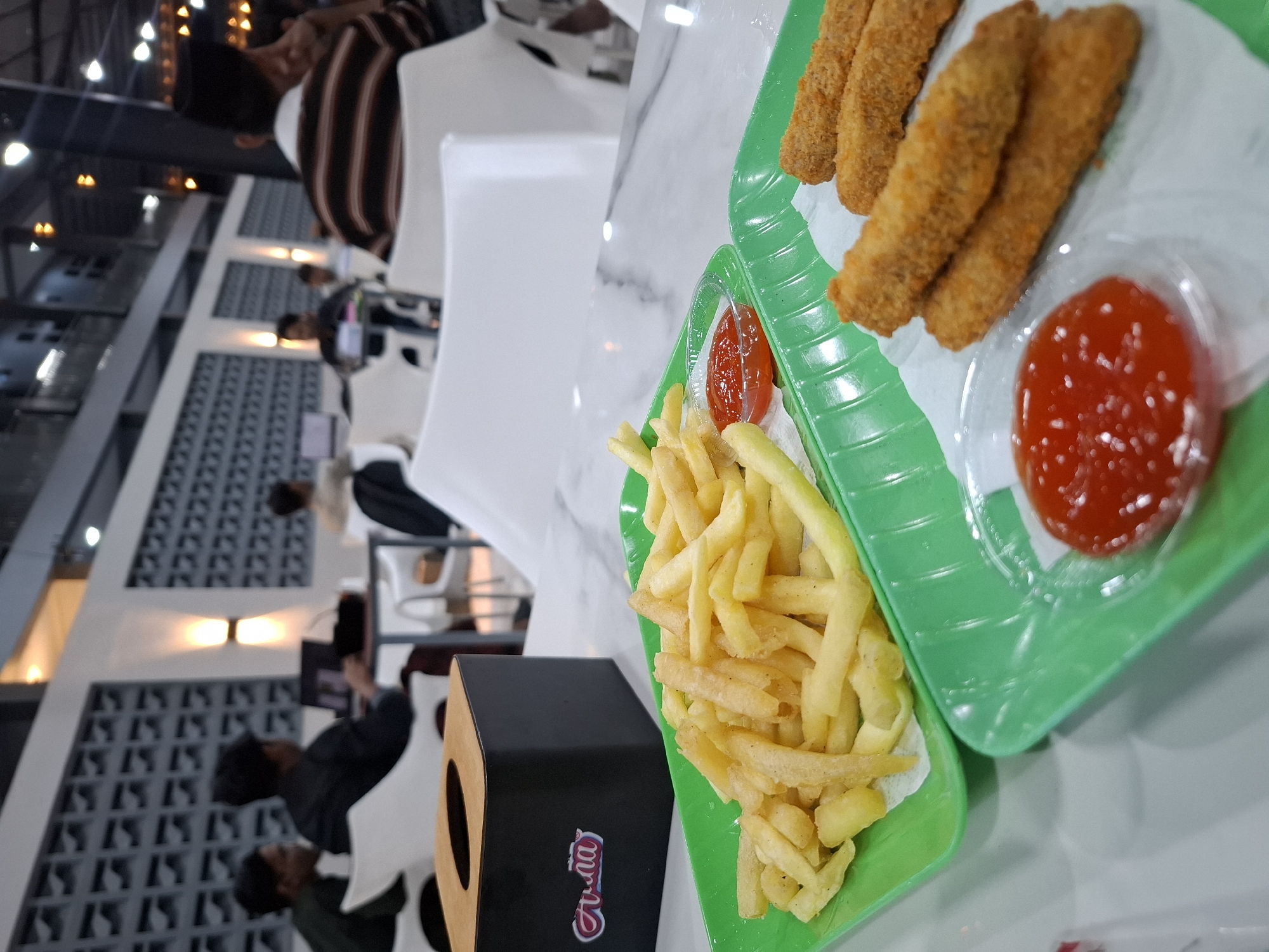 In Kampongbaro, Indonesia, a dimly lit establishment displays two green plastic trays of food on a white marble-patterned table. The tray in the foreground contains several breaded chicken tenders and a small dish of ketchup. To the left, a second green tray is filled with golden french fries, also accompanied by a small dish of ketchup. A black tissue box with a wooden lid is positioned behind the food, bearing a red and white logo. In the background, several individuals are seated at tables, some appearing to be working on laptops. The environment suggests a casual dining or co-working space, with white chairs and grey decorative wall panels visible.