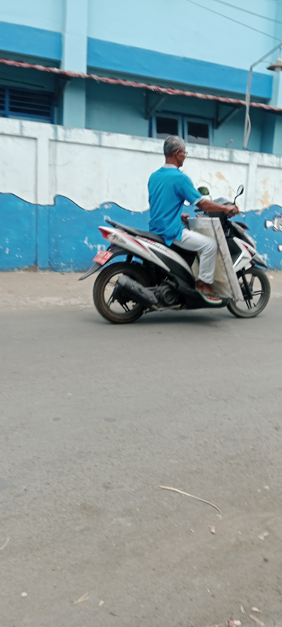 An elderly man with graying hair and glasses is riding a white and black scooter on a paved road in what appears to be Purwomartani, Indonesia. He is wearing a blue short-sleeved shirt and light-colored pants. The scooter has a dark-colored exhaust pipe and a license plate with red lettering. A light-colored fabric, possibly a mat or cover, is draped over the front of the scooter. Behind the scooter, a building with blue and white painted walls is visible. The blue sections of the building have a wavy pattern, and there are windows with blue frames. The road surface is asphalt and shows signs of wear. The overall scene depicts a typical street view in a moderately populated area.