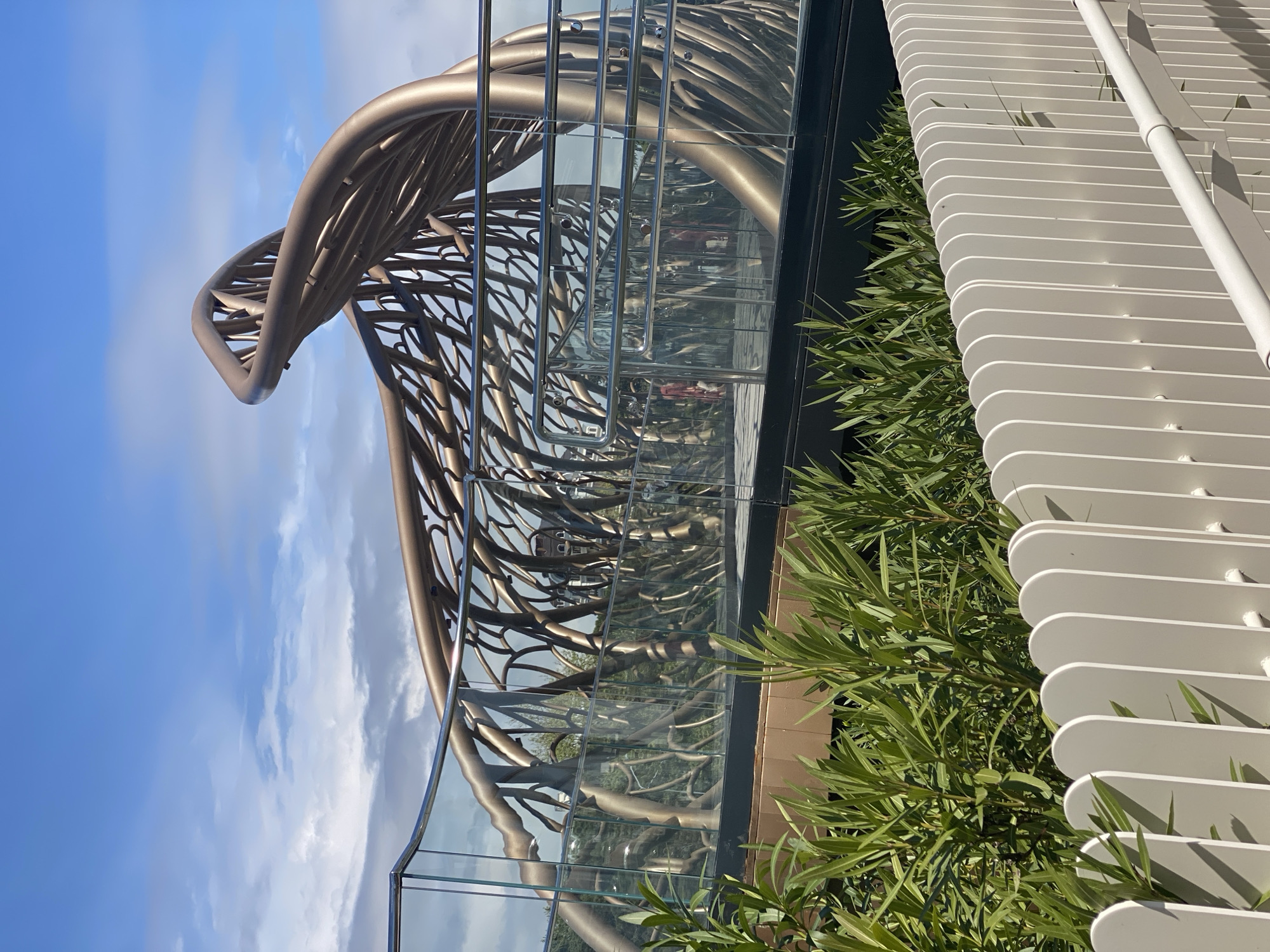 The image features an architectural structure, possibly a pedestrian bridge or a viewing platform, notable for its complex design of intertwined, tubular elements in a metallic bronze finish. Clear glass panels form a barrier along the edge of the structure. The background reveals a blue sky with scattered clouds.
To the right of the structure, there's a series of closely spaced, white, rounded architectural louvers or screens. Green foliage of shrubs or trees is visible, intermingling with the louvers and adjacent to the structure. Reflections on the glass panels suggest urban surroundings.
Based on the design and architecture, the location is highly suggestive of the "DNA Bridge" in Sochi, Russia, known for its distinctive double helix-inspired form.